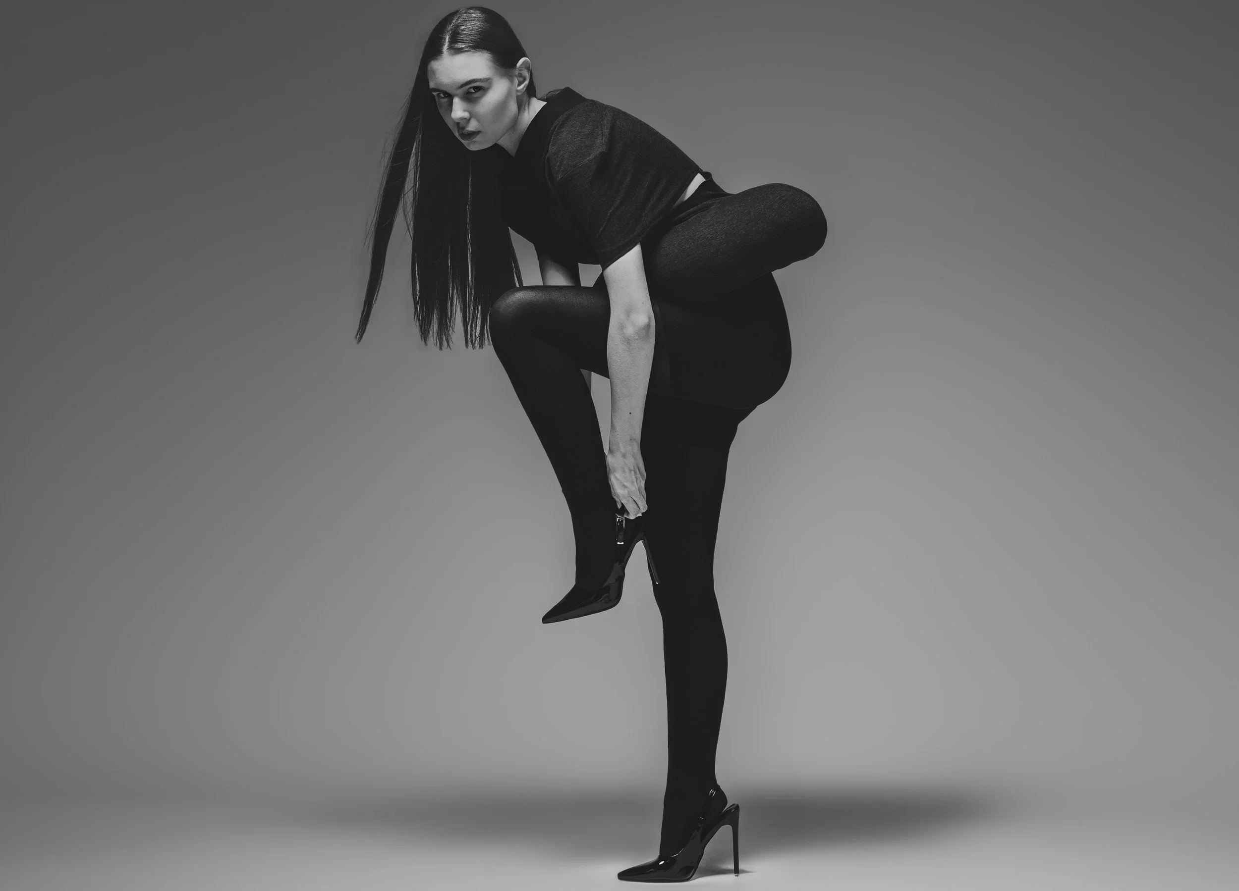 Femme en tenue noire portant des talons hauts, posant contre un fond gris.