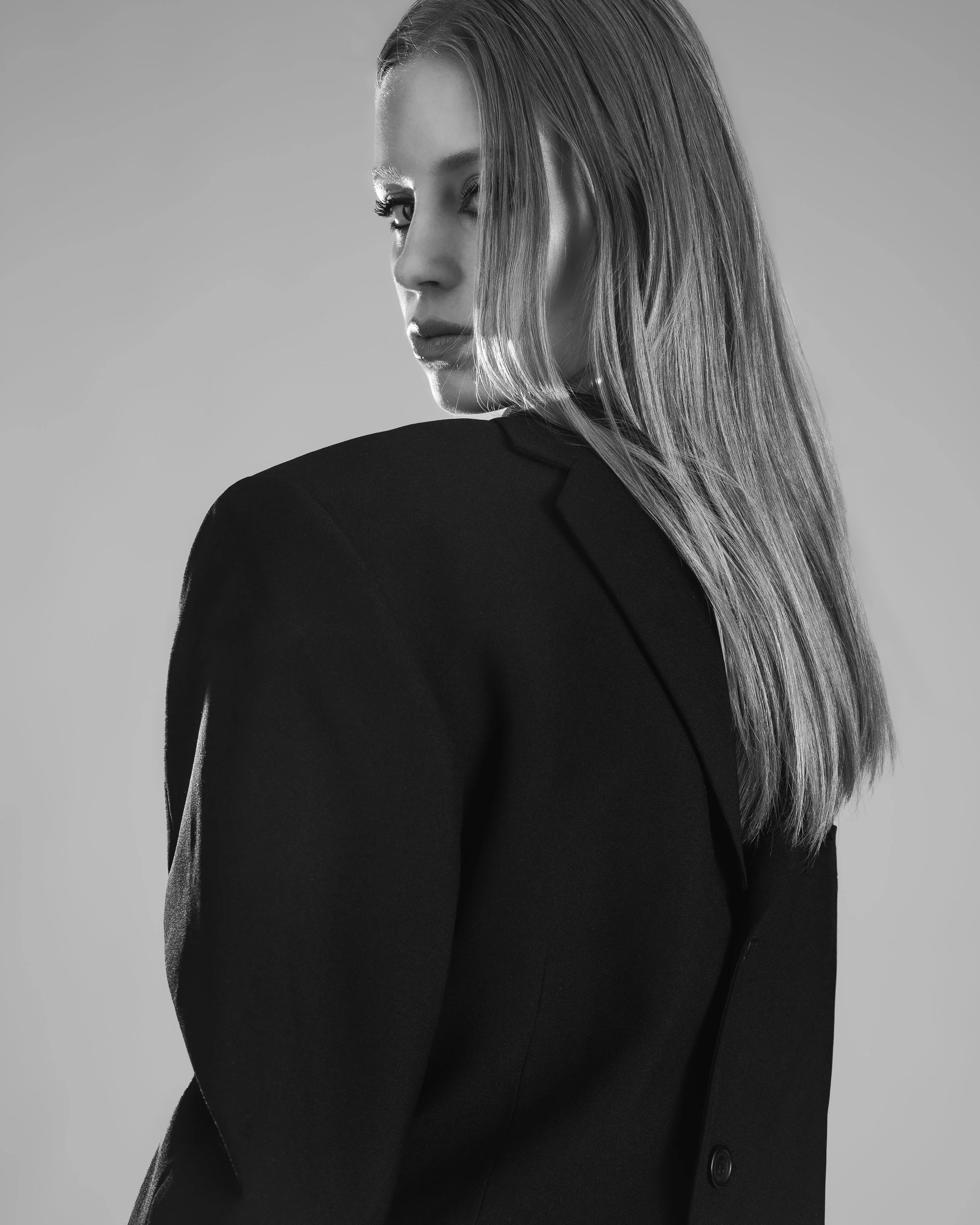 Une femme avec des cheveux longs et lisses porte un blazer noir, vue de dos avec un léger regard sur le côté, en noir et blanc.