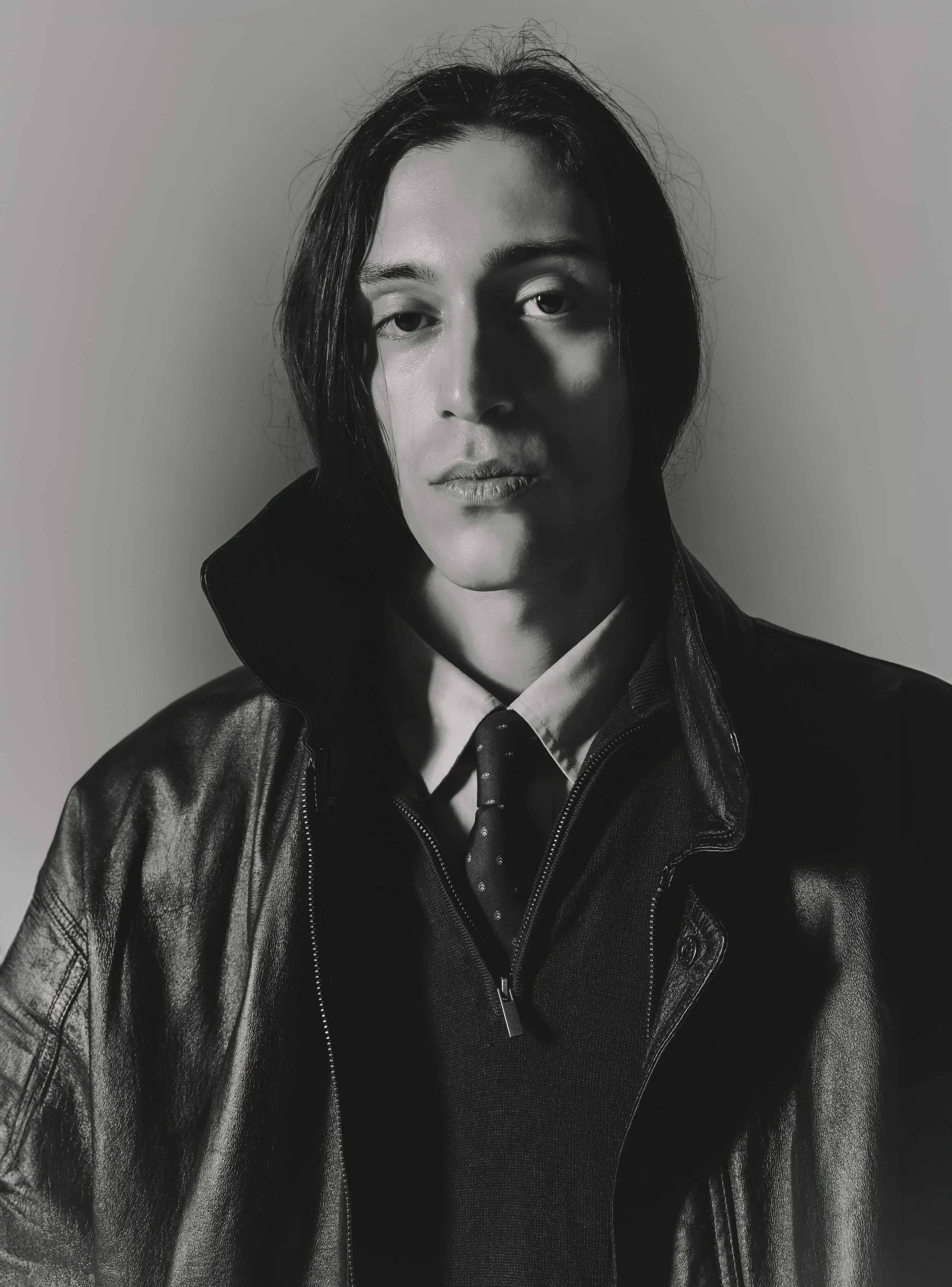 Un homme avec des cheveux longs, portant une veste en cuir, une chemise et une cravate à pois, regardant vers l'objectif, en noir et blanc.