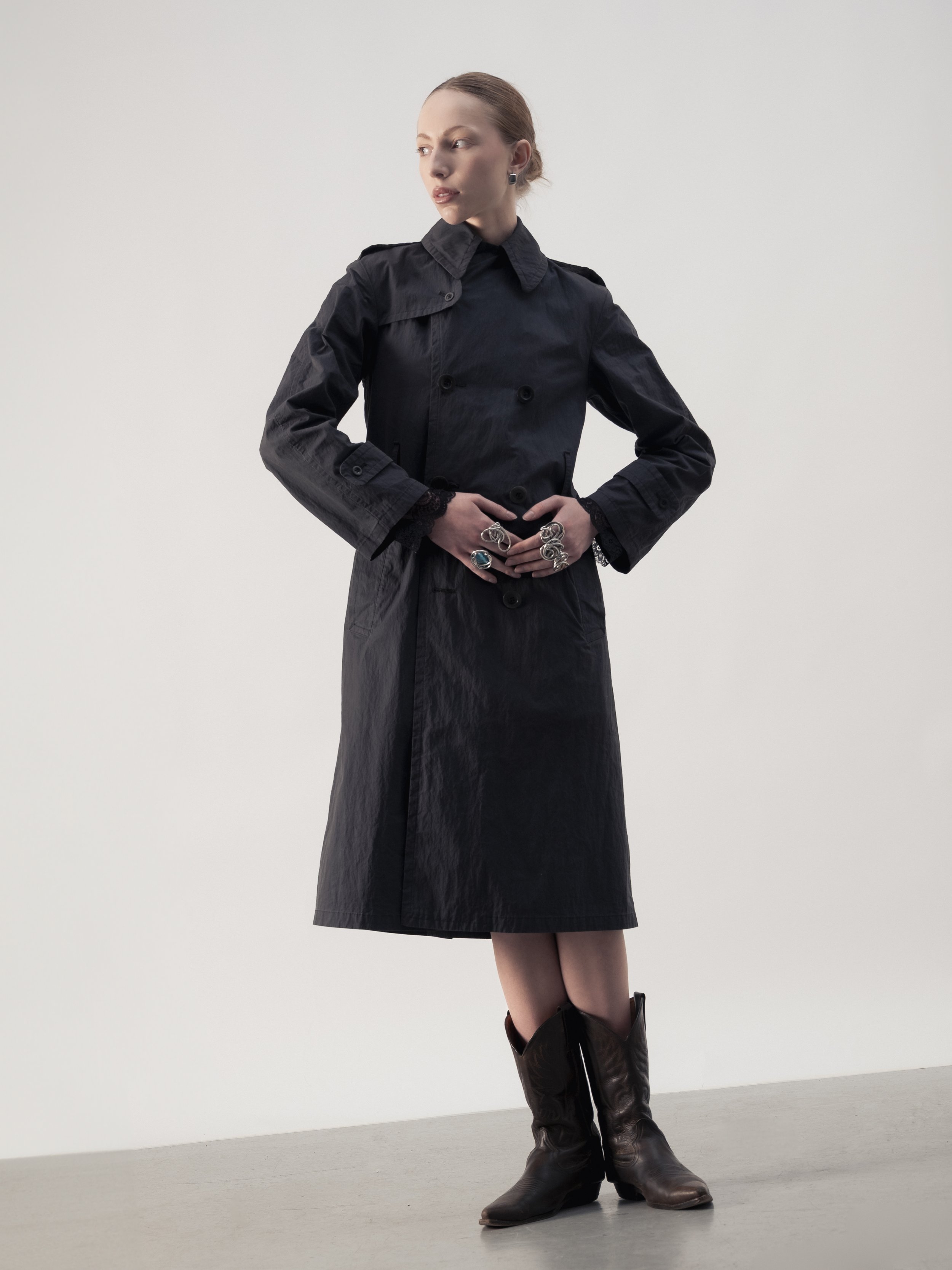 Une femme porte un trench noir, des bottes en cuir brunes, et plusieurs bagues, se tenant contre un fond blanc.