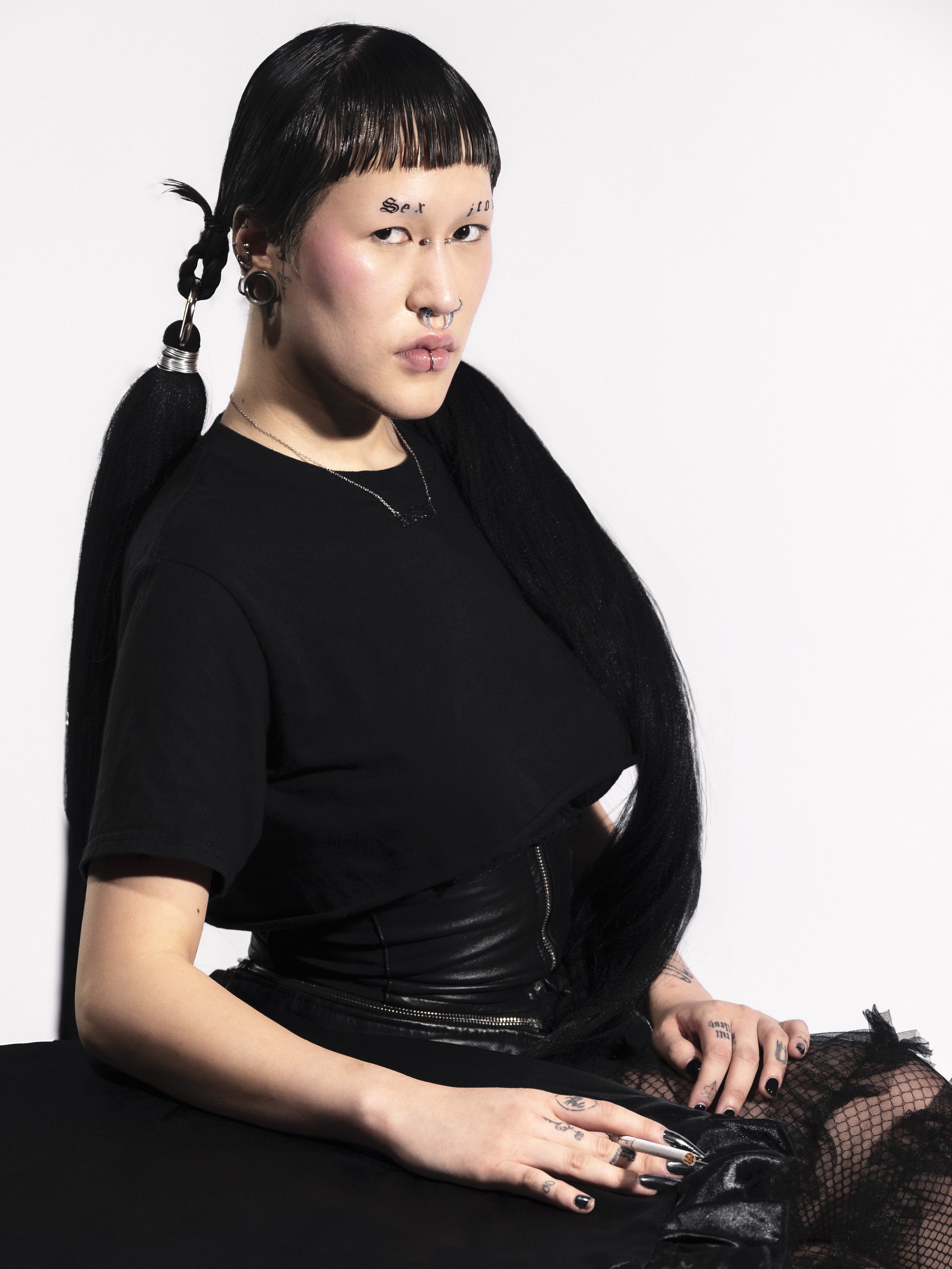 Jeune femme asiatique avec des piercings, des tatouages et des cheveux noirs, portant un t-shirt noir et des vêtements noirs, assise contre un fond blanc.