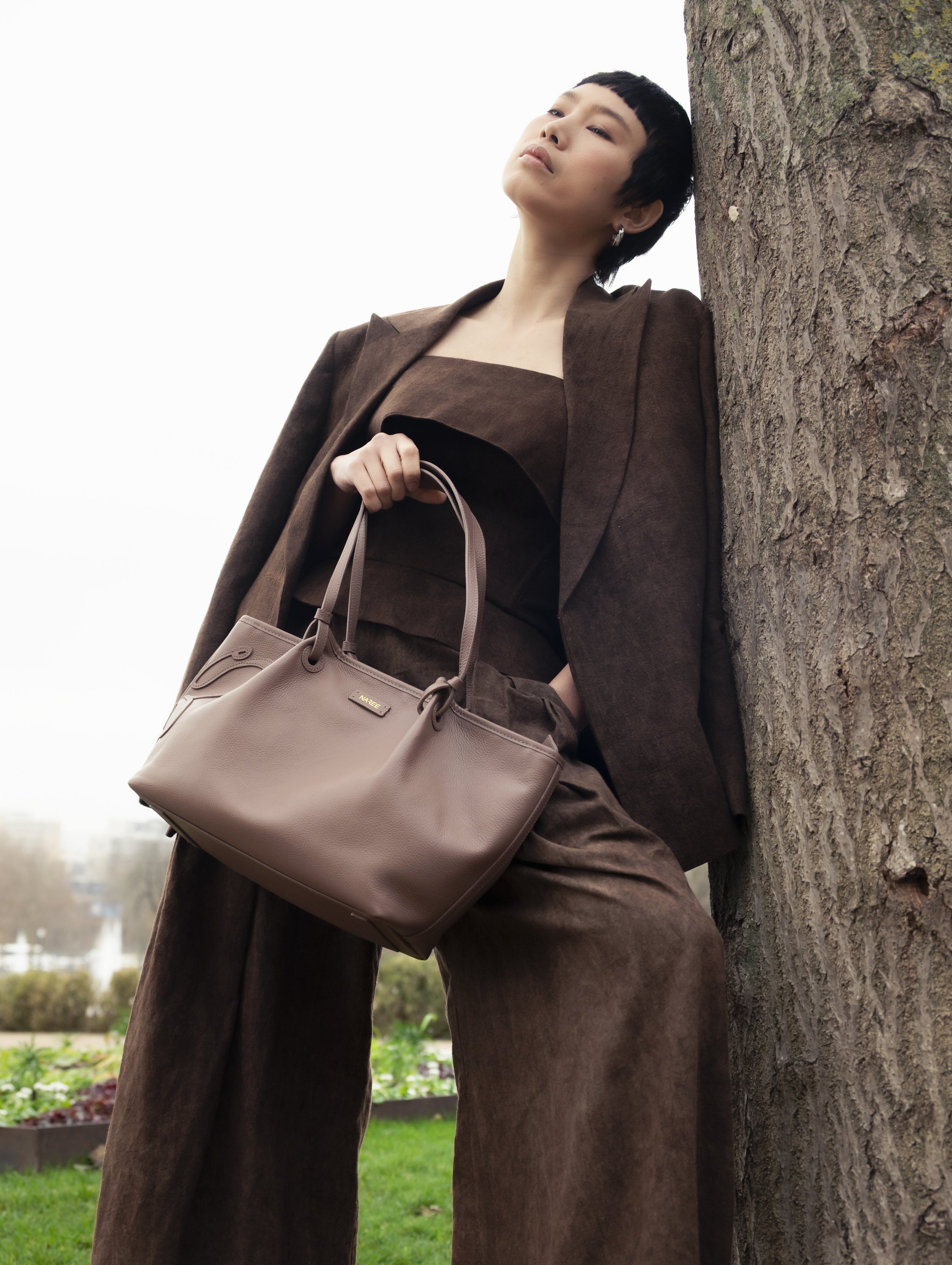 Une femme élégante avec un tailleur marron, portant un sac à main beige, se tient contre un arbre dans un parc.