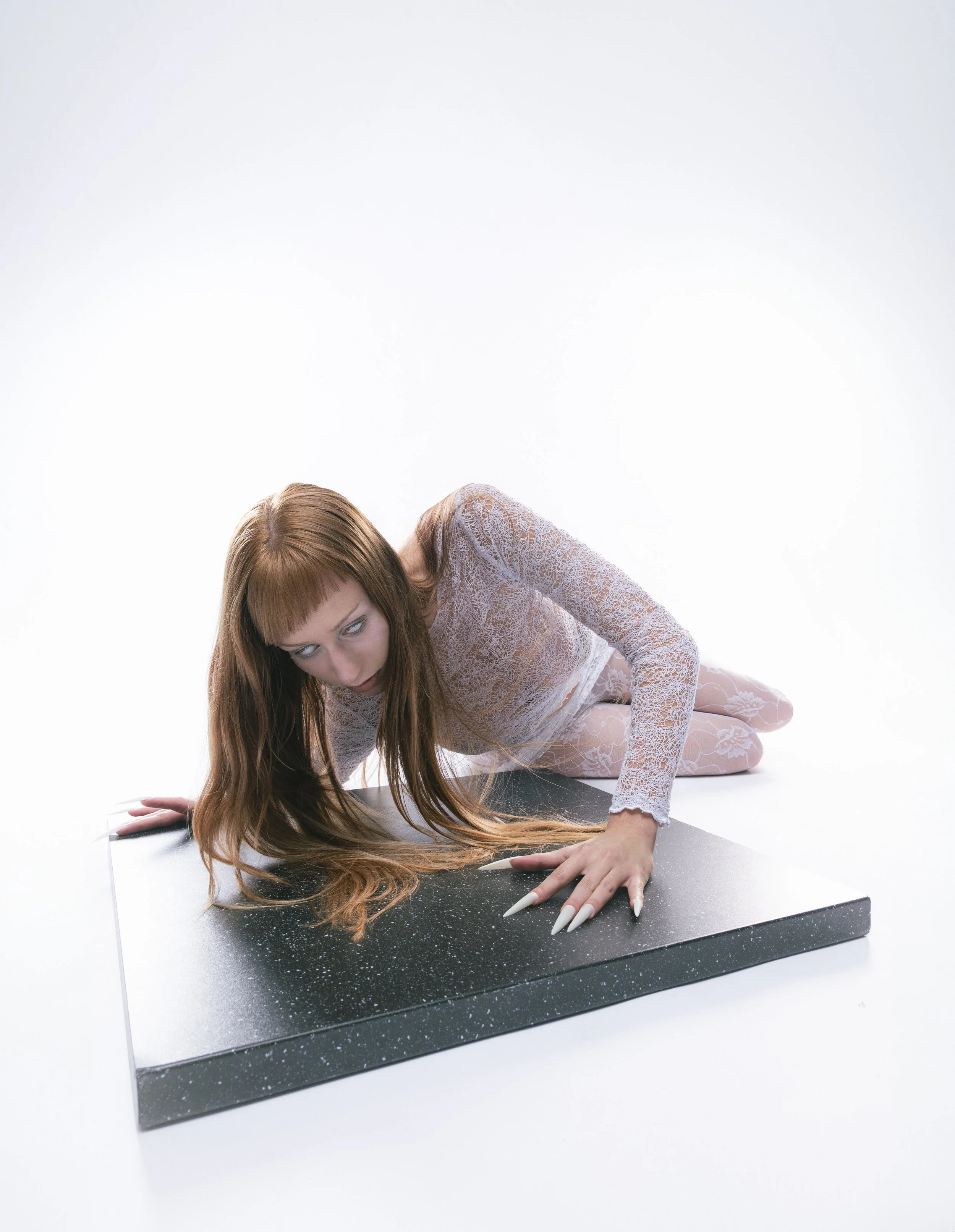 Femme jeune aux cheveux roux, habillée d'une robe en dentelle blanche, allongée sur un plateau noir avec des détails blancs, dans un fond blanc.