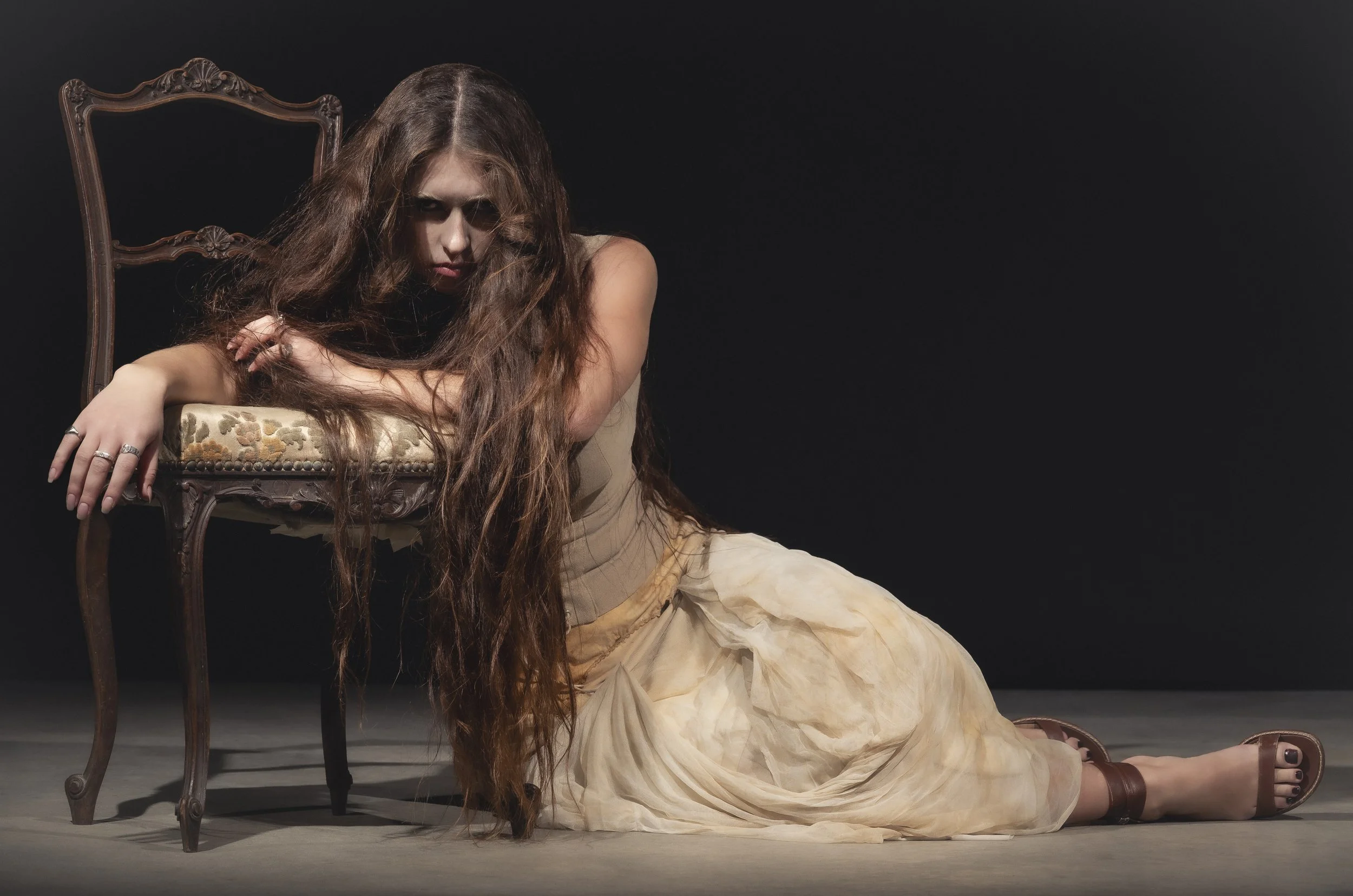 Une jeune femme aux longs cheveux bruns, vêtue d'une robe beige claire, est allongée sur le sol, appuyée sur une chaise antique en bois avec un coussin beige. Son regard est intense et elle porte plusieurs bagues à ses doigts. Le fond est noir, créan