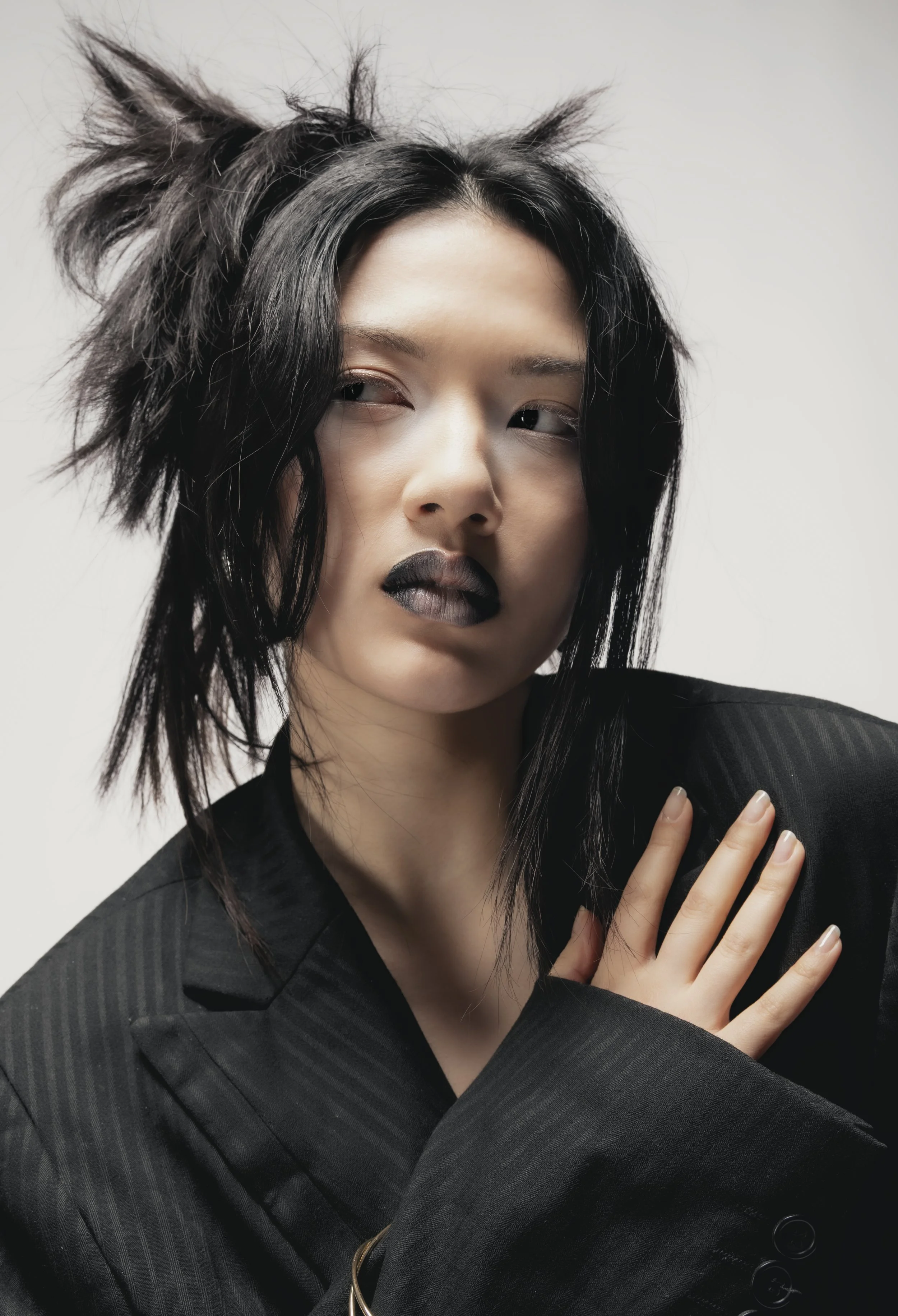Un portrait d'une femme avec des cheveux noirs en désordre, portant un maquillage sombre, vêtue d'un costume noir avec un fond clair.
