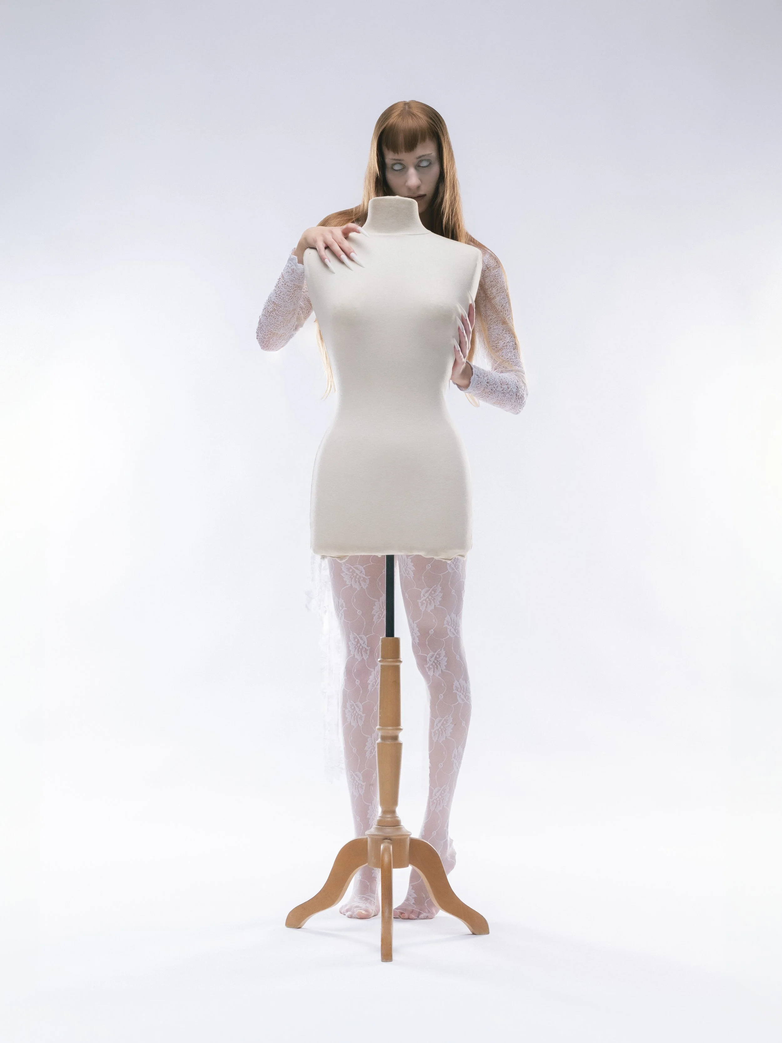 Une femme avec des yeux blancs, portant des vêtements en dentelle blanche, se tient derrière un mannequin de couture blanc sur un fond neutre.