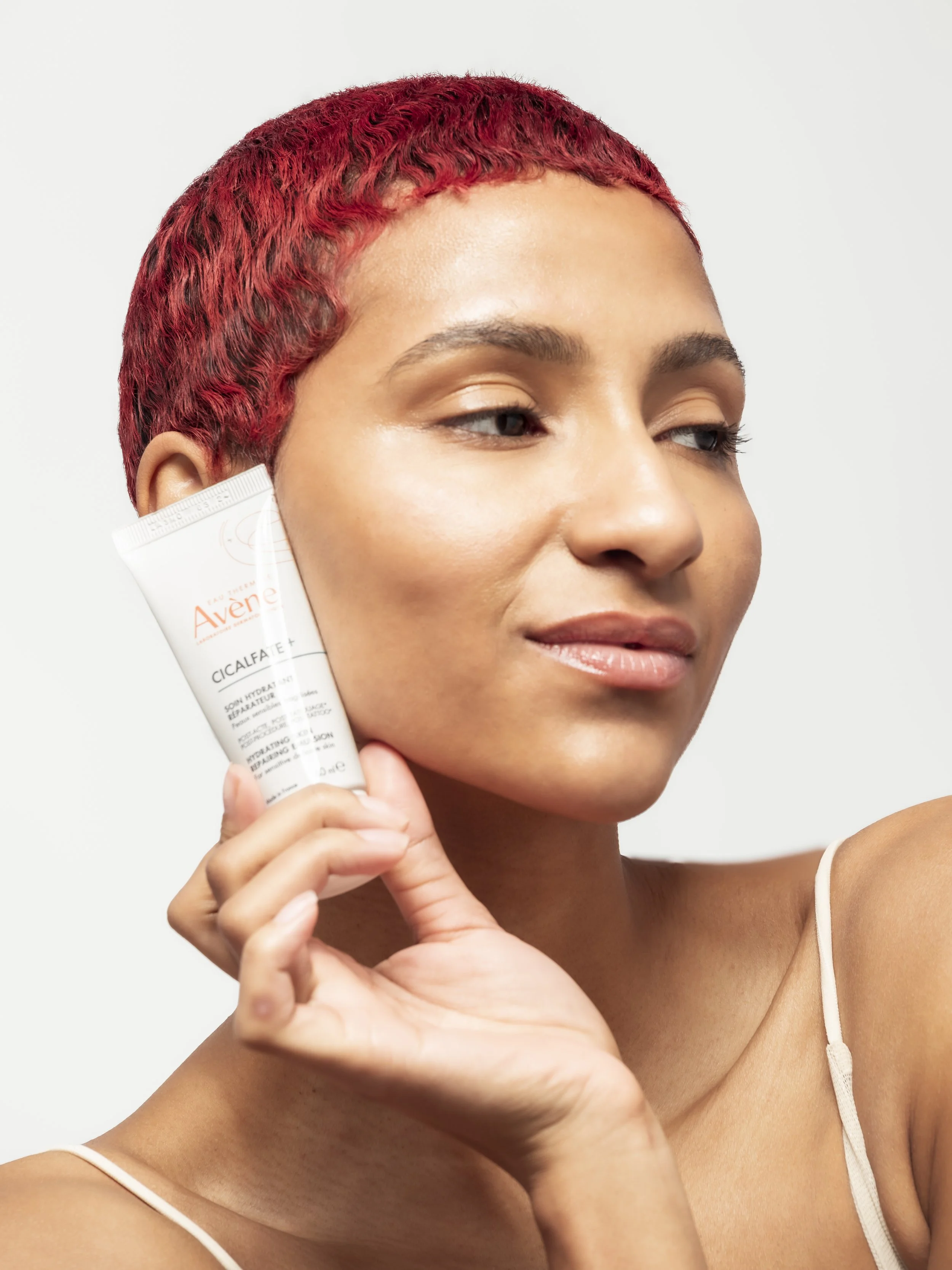 Jeune femme avec cheveux courts et roux, tenant un tube de crème de marque Avène près de l'oreille.