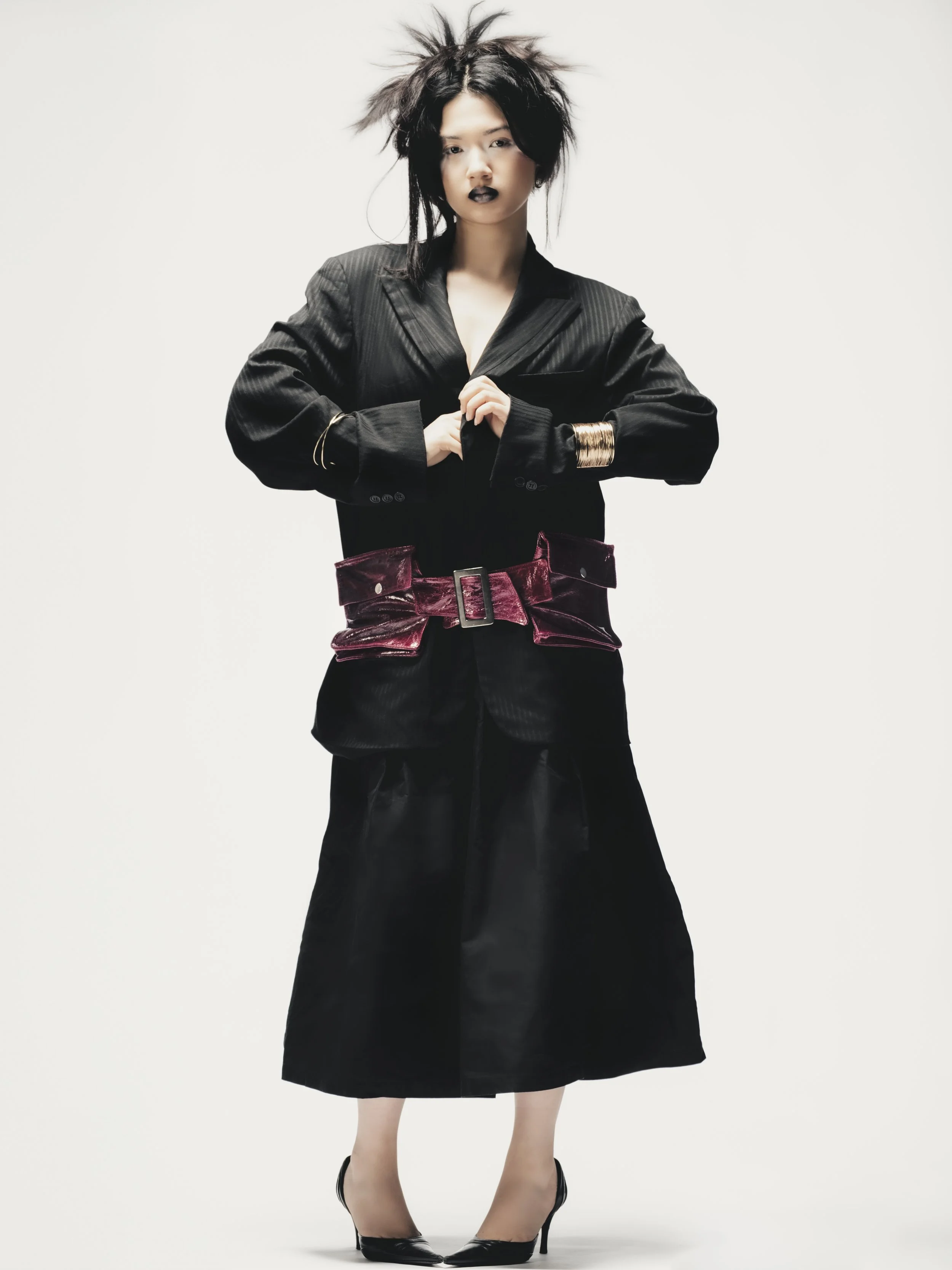 Femme portant un costume noir avec une large ceinture en velours rouge, cheveux en désordre, maquillage sombre, debout contre un fond blanc.