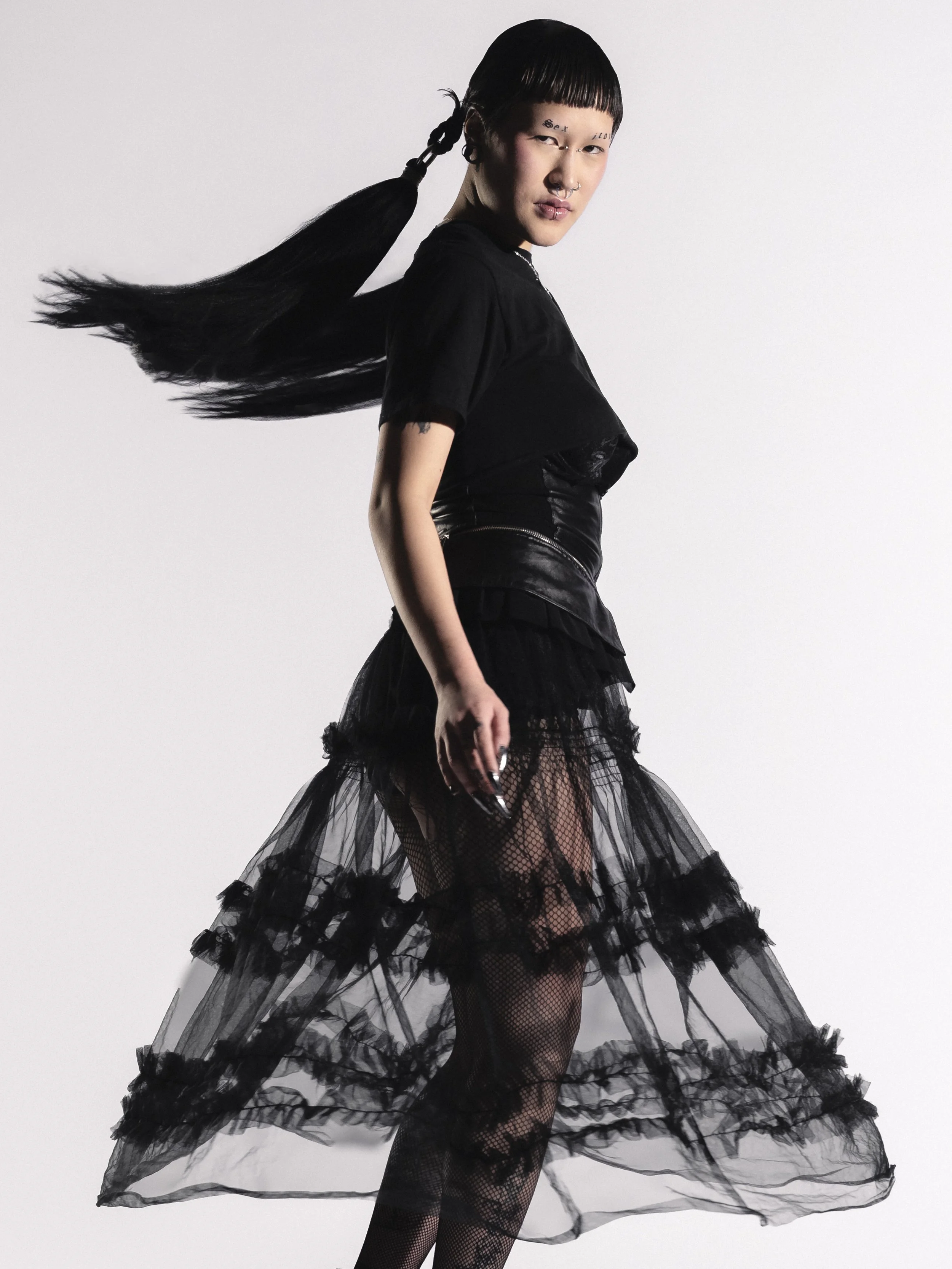 Une femme portant une tenue noire avec une jupe en tulle transparent et des détails en satin, pose sur un fond blanc.