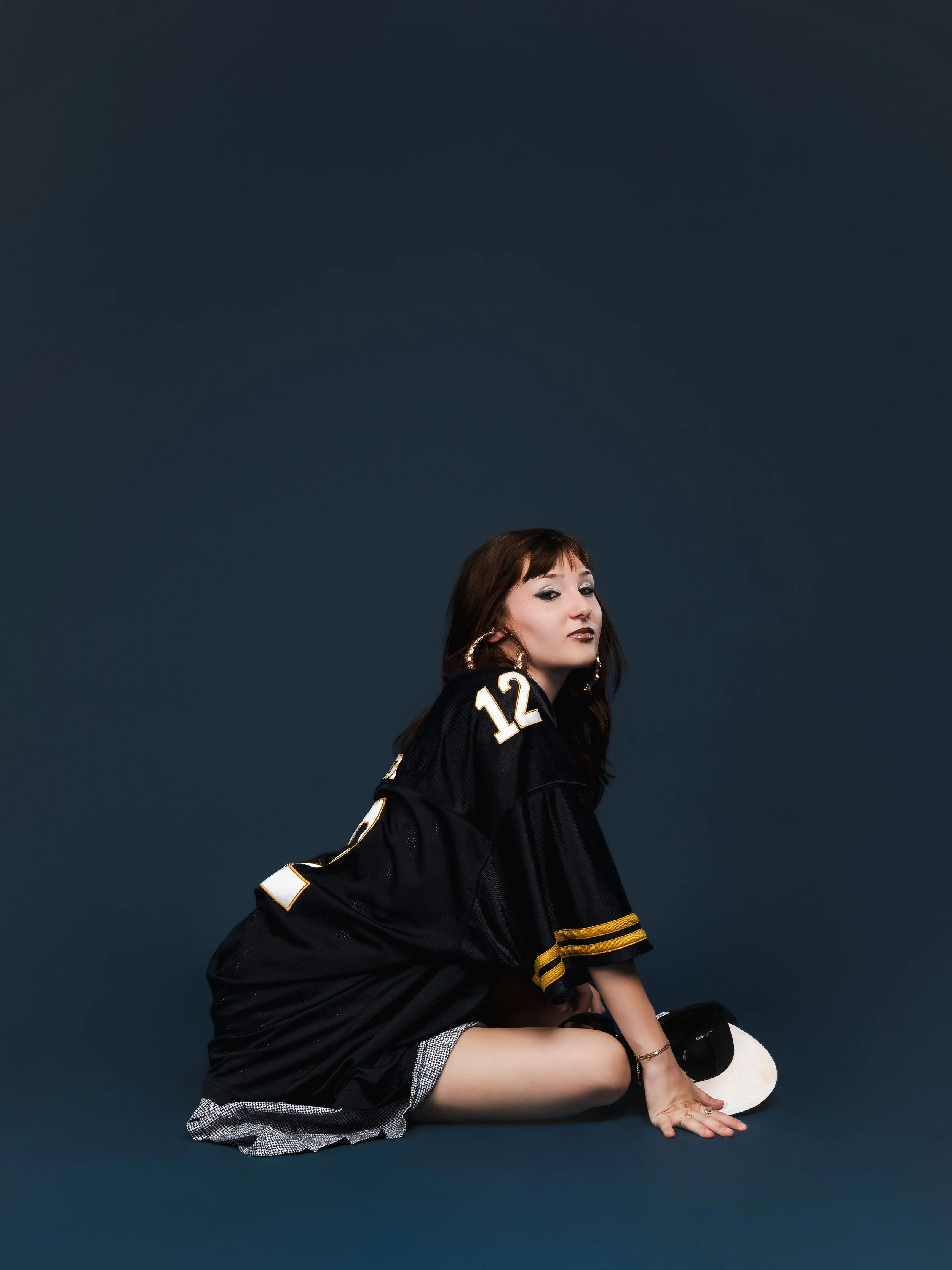 Une femme avec des cheveux bruns, portant un maillot de football noir avec le numéro 12, assise sur le sol avec une main au sol, avec un chapeau noir et blanc à côté, contre un fond gris foncé.