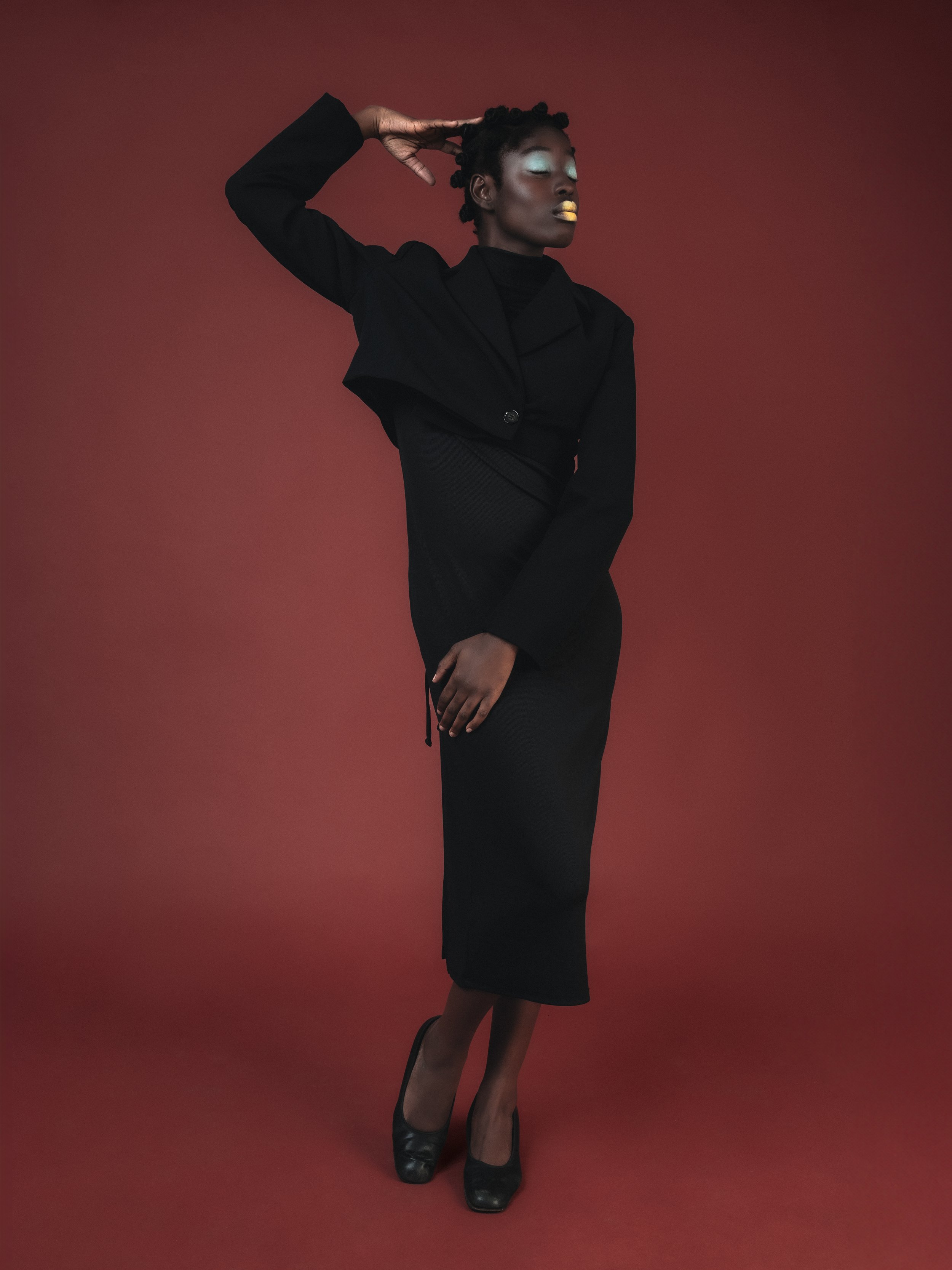 Une femme noire élégante portant un tailleur noir, posant avec une main derrière la tête et l'autre posée sur la hanche, avec un fond rouge.