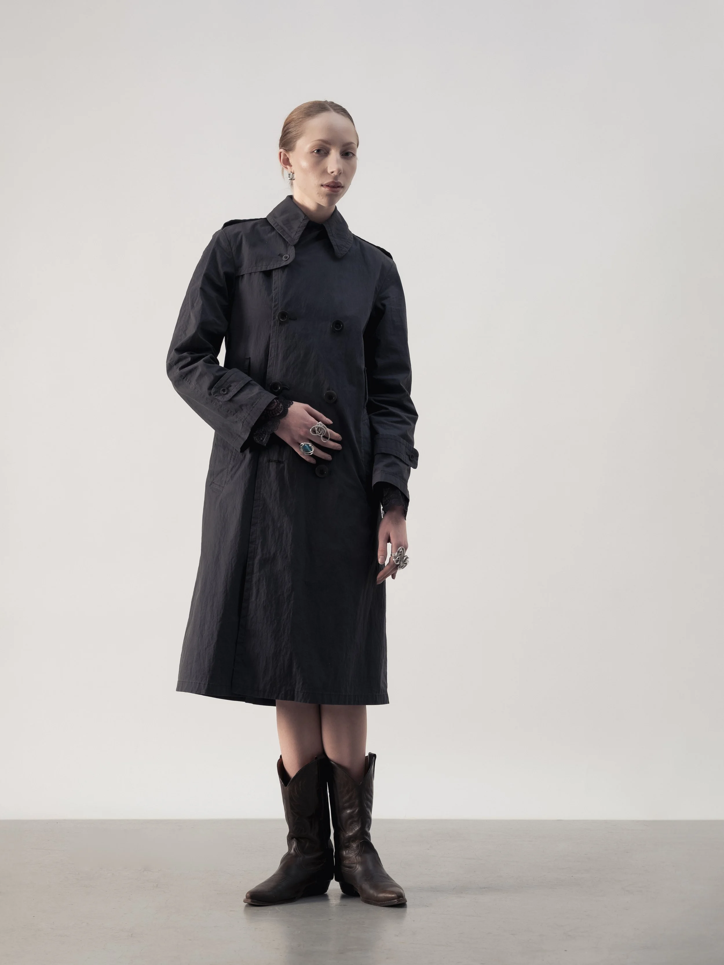 Une femme portant un trench noir, des bottes en cuir marron, et de multiples bagues visibles.
