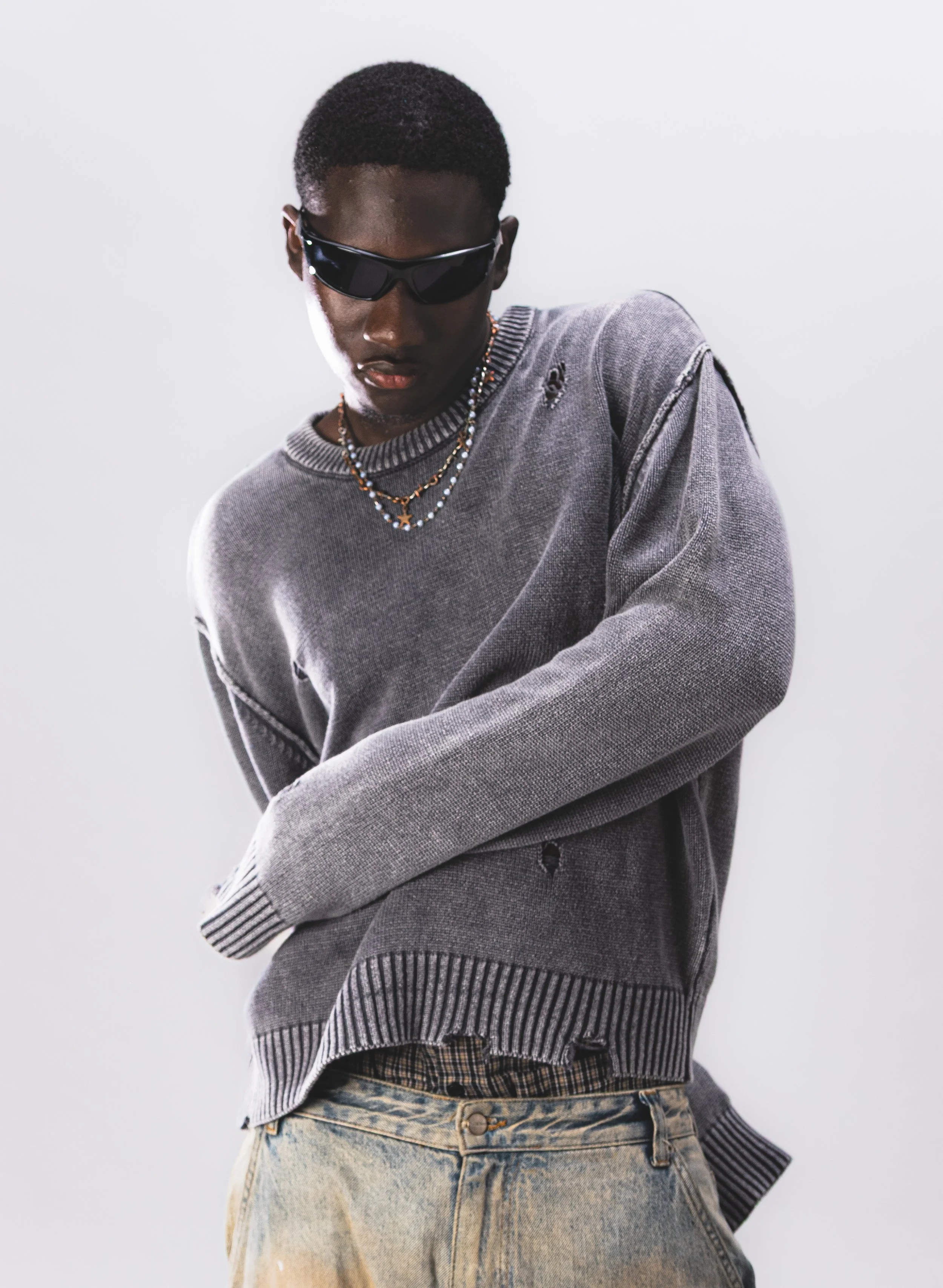 Jeune homme portant des lunettes de soleil noires, un sweat gris déchiré, un jean clair et des colliers, posé contre un fond blanc.