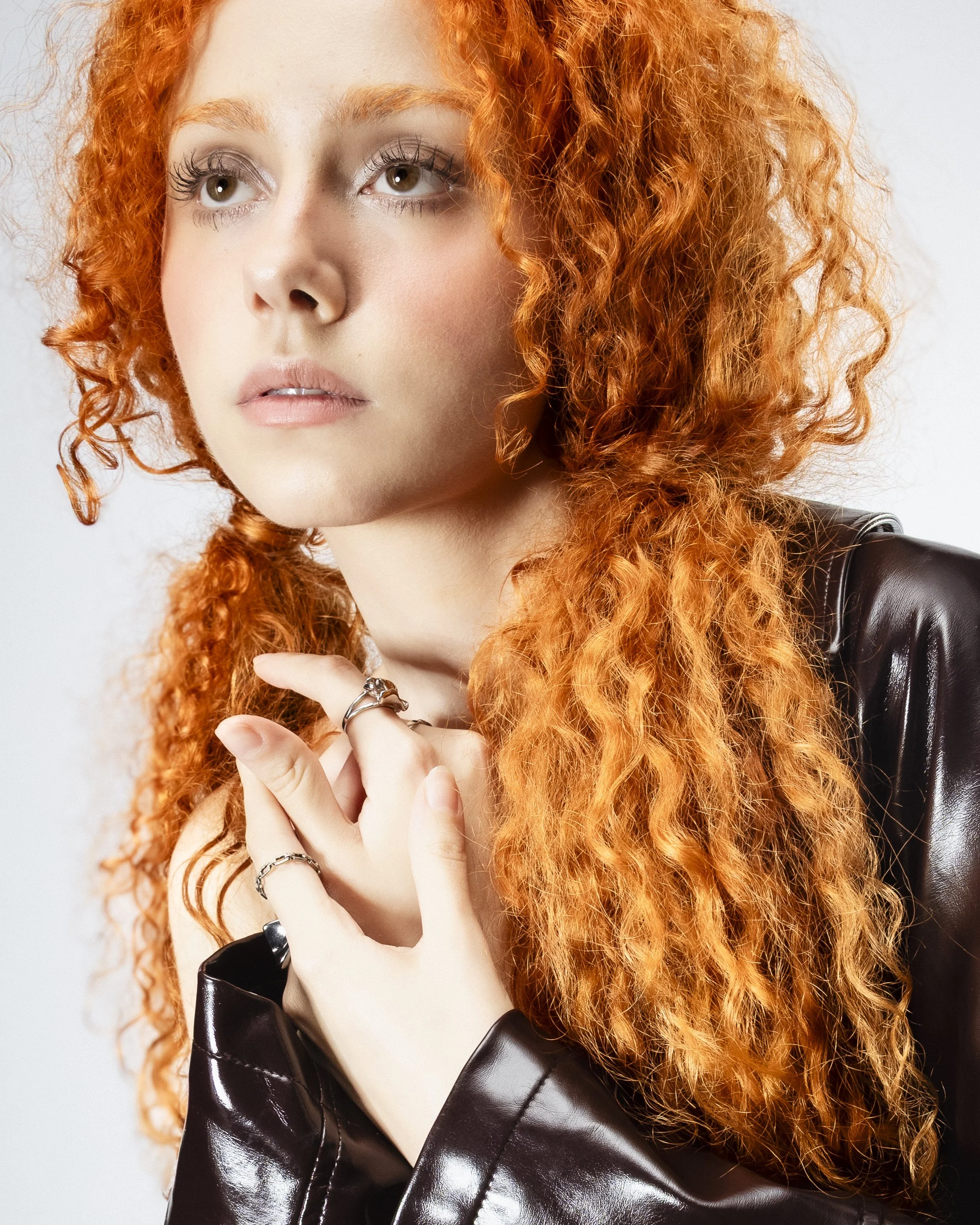 Jeune femme avec cheveux roux bouclés, portant un manteau en cuir noir, regardant vers la gauche, mains avec des bagues, fond gris clair.
