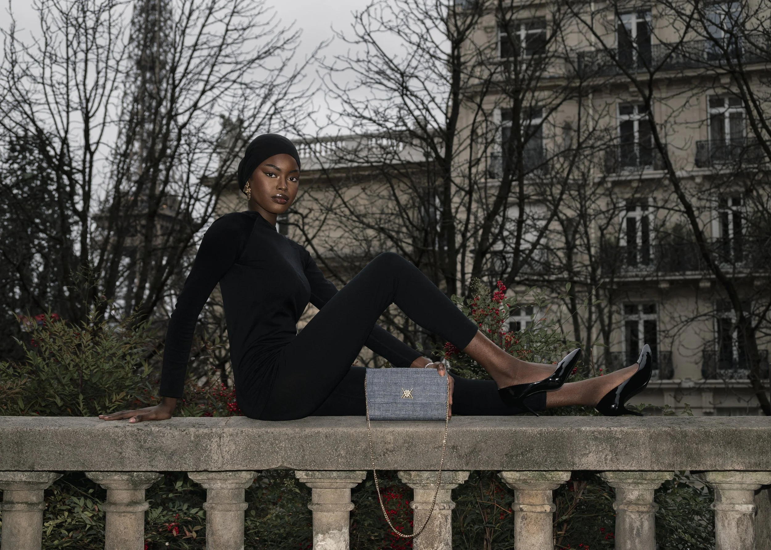 Une femme élégante en tenue noire, portant un pantalon, un haut à manches longues, des talons noirs, et un foulard noir, est assise sur une balustrade en pierre dans un environnement urbain avec des bâtiments en arrière-plan, des arbres sans feuilles