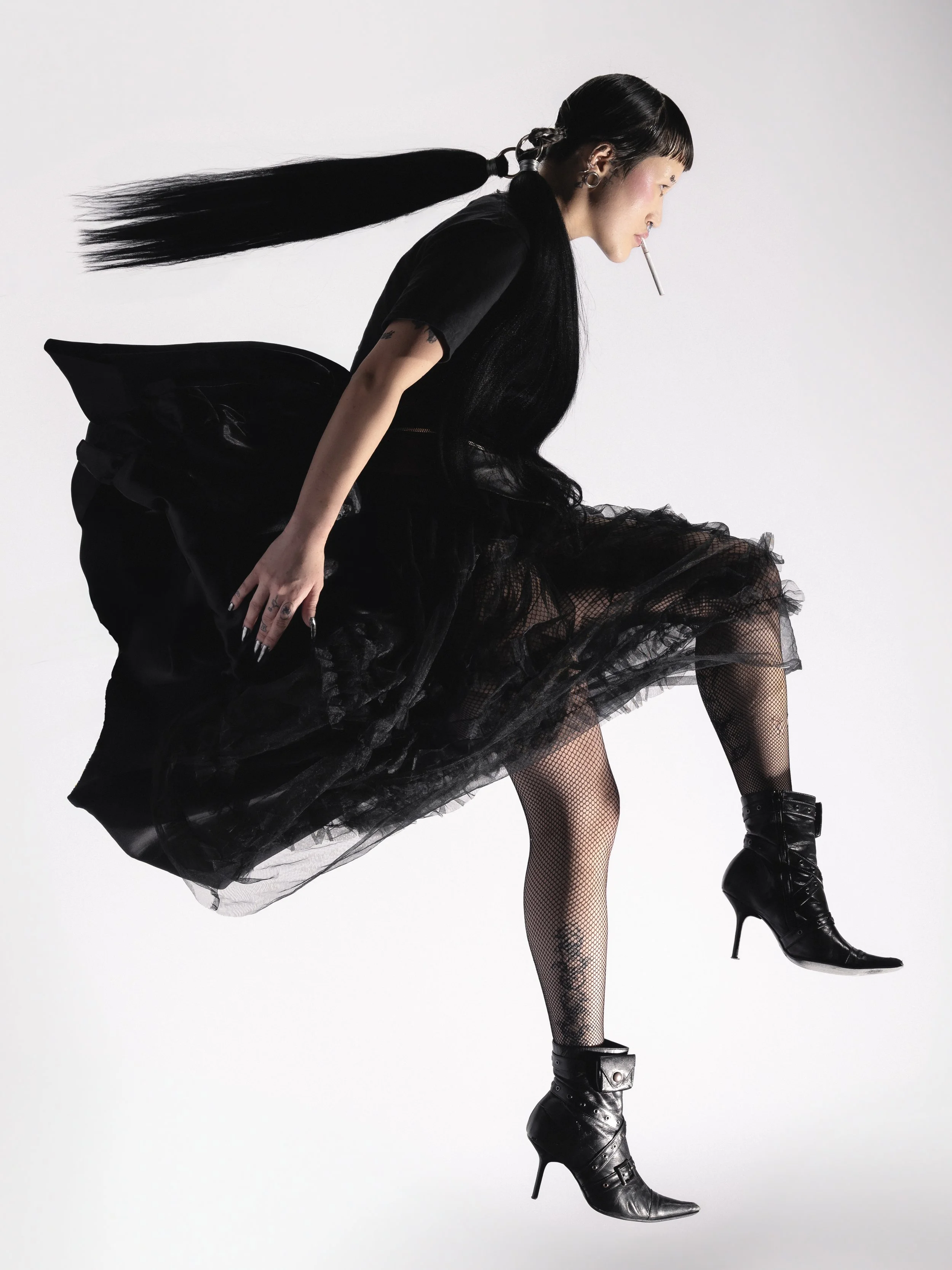 Une femme habillée en noir avec des talons aiguilles noirs, portant une robe en tulle noire, avec des vêtements en dentelle et un piercing à la lèvres, tient une cigarette dans la bouche.