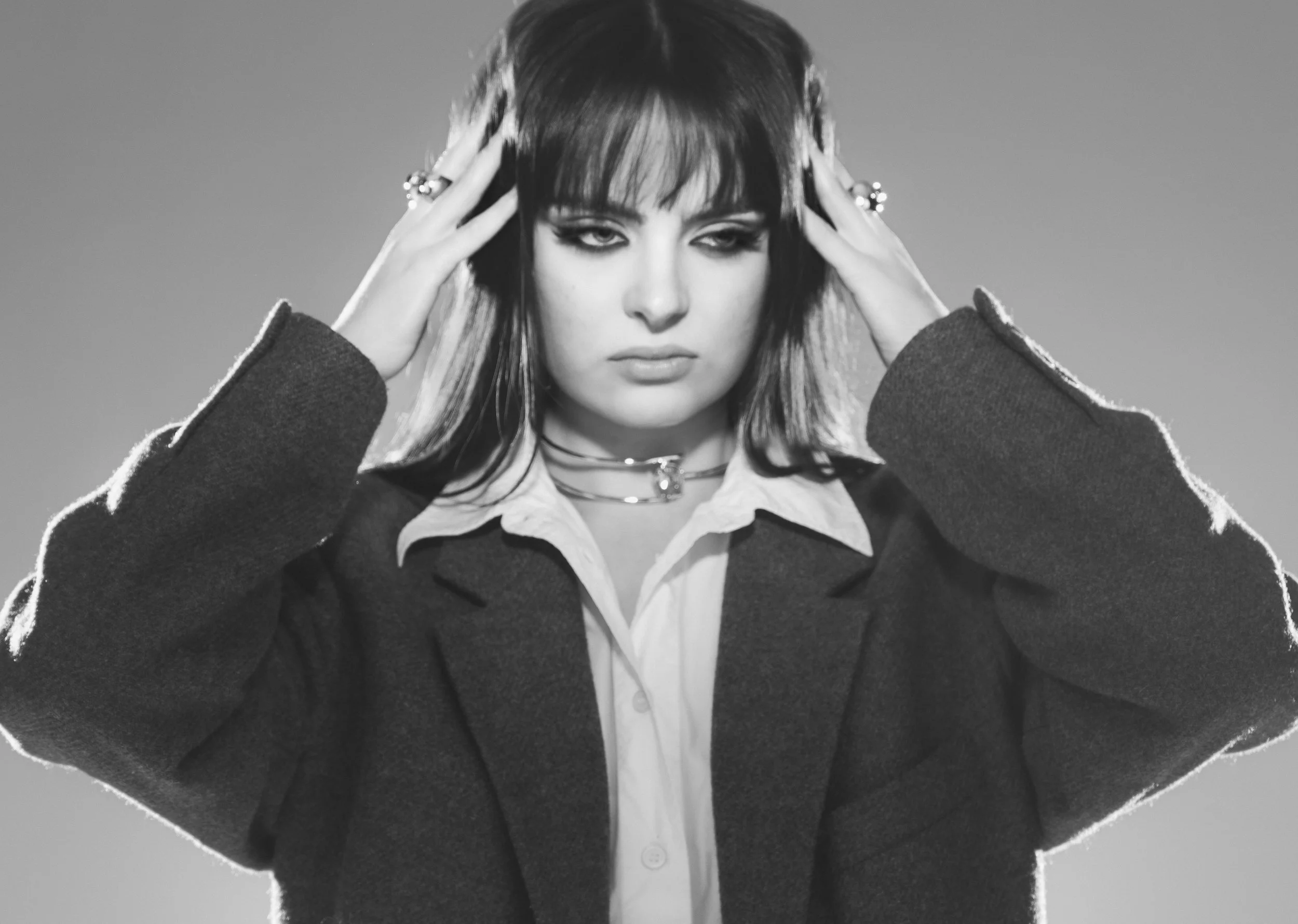 Une femme en noir et blanc, portant une veste foncée et une chemise claire, avec des cheveux courts et coiffés en bataille, tient ses mains à la tête, exprimant une émotion intense.