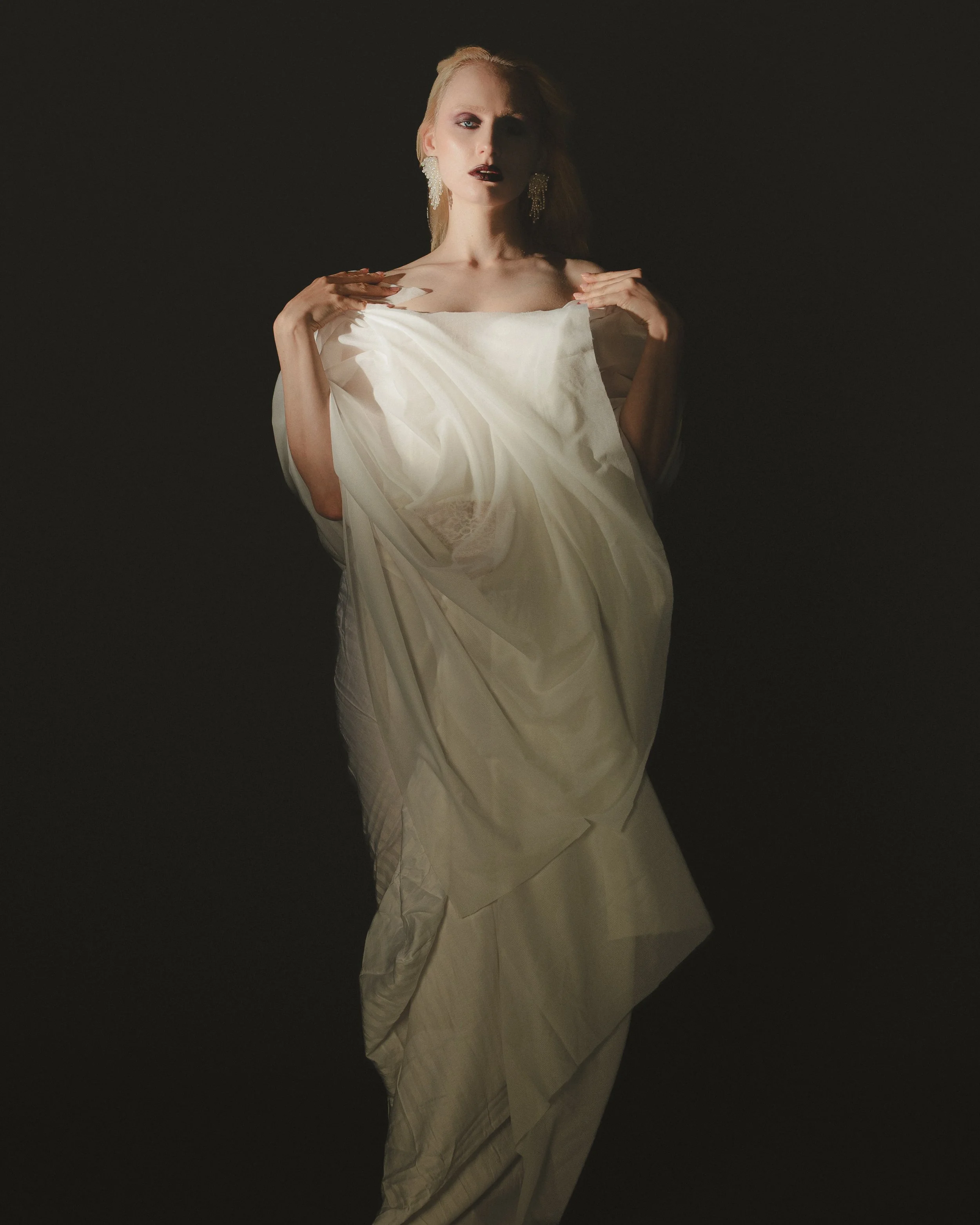 Une femme porte une robe de mariée blanche avec des boucles d'oreilles pendantes, dans un éclairage sombre.