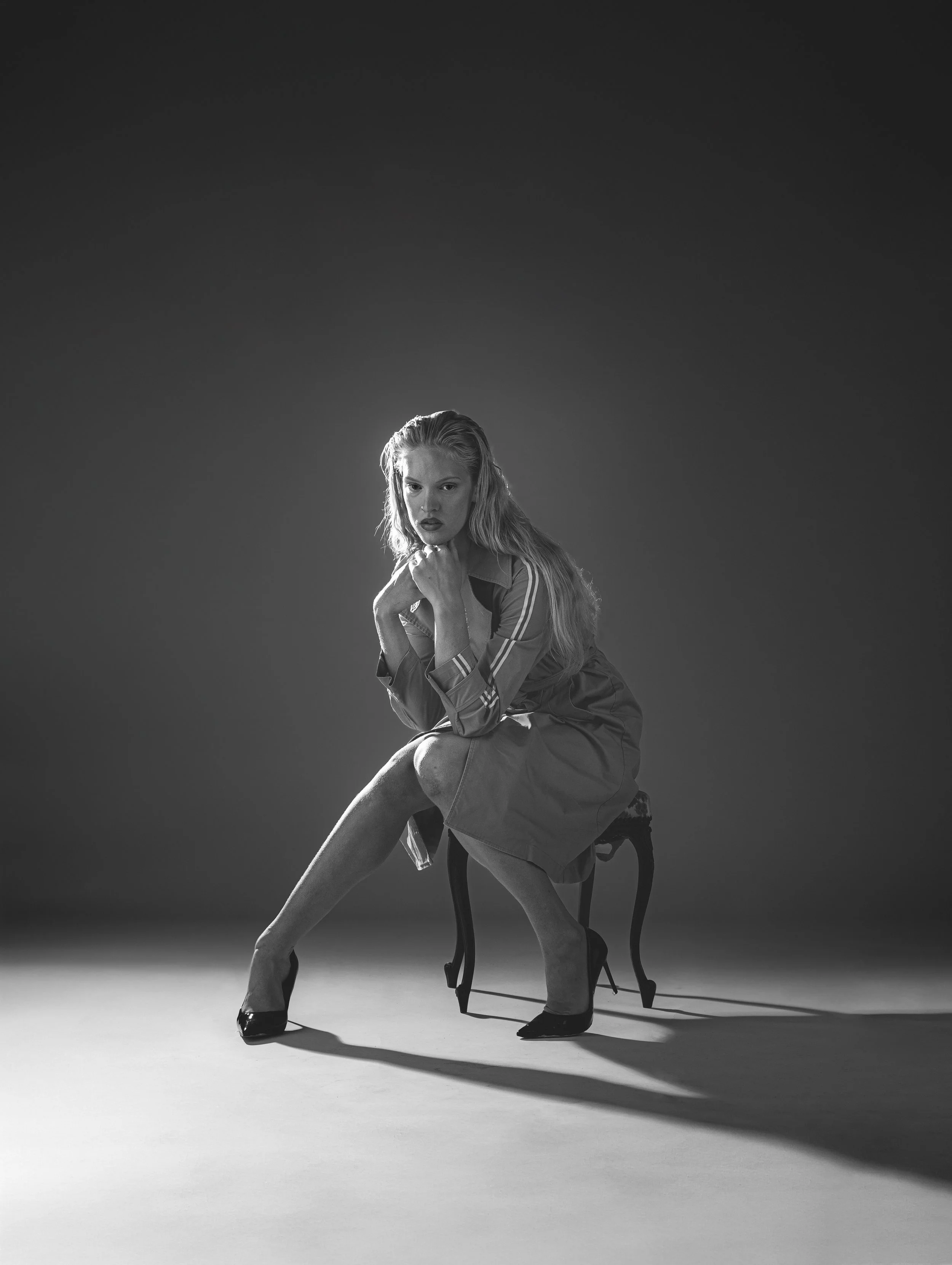 Femme en noir et blanc, assise sur une petite chaise, portant une robe, chaussures à talons, dans un studio avec un éclairage dramatique.