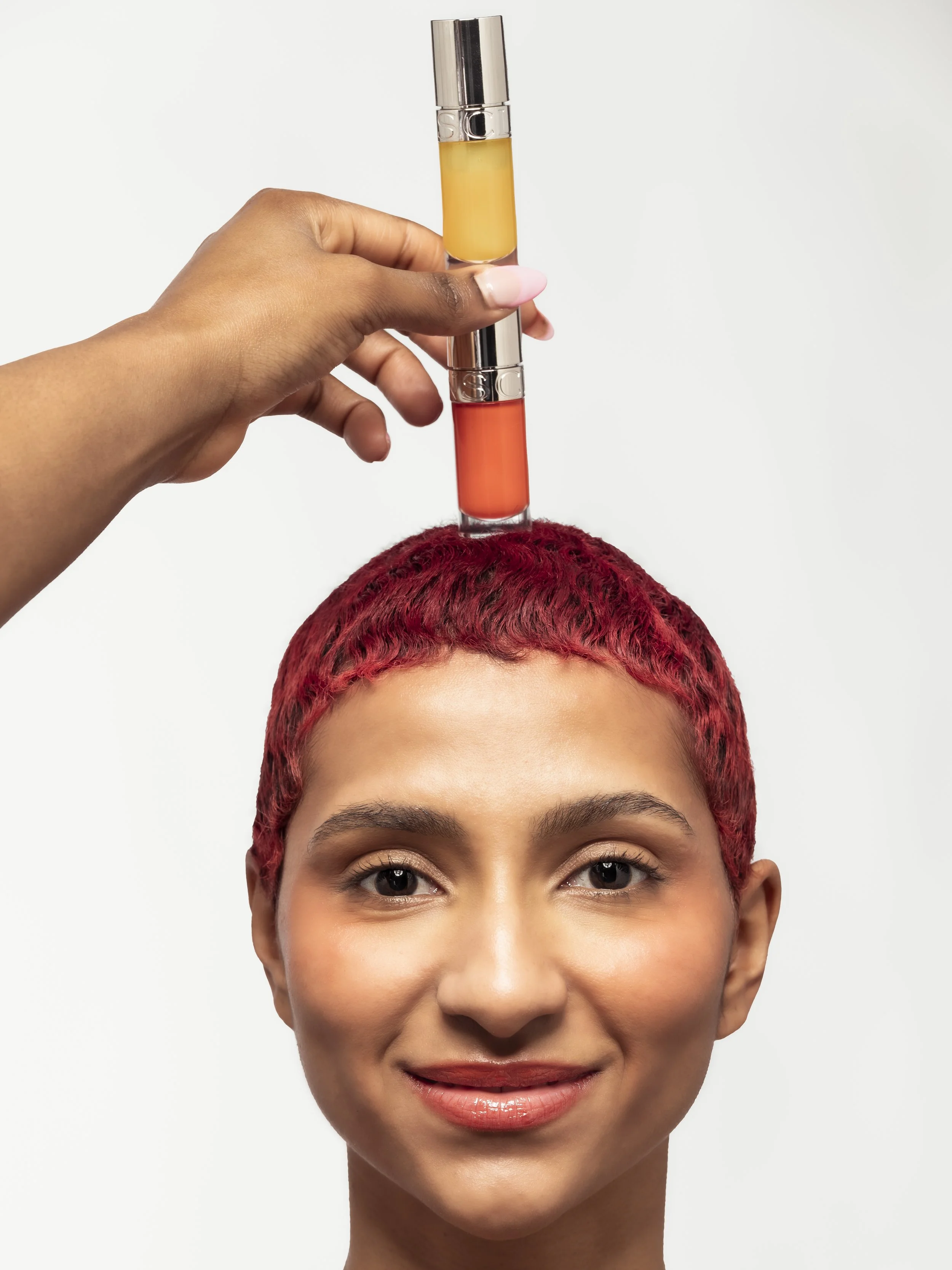Une femme à la peau claire et aux cheveux courts teints en rouge, souriante, regarde devant elle. Une main tient un test de tubulure contenant deux sections de couleurs différentes, rouge en bas et jaune en haut, équilibrées au sommet de sa tête.