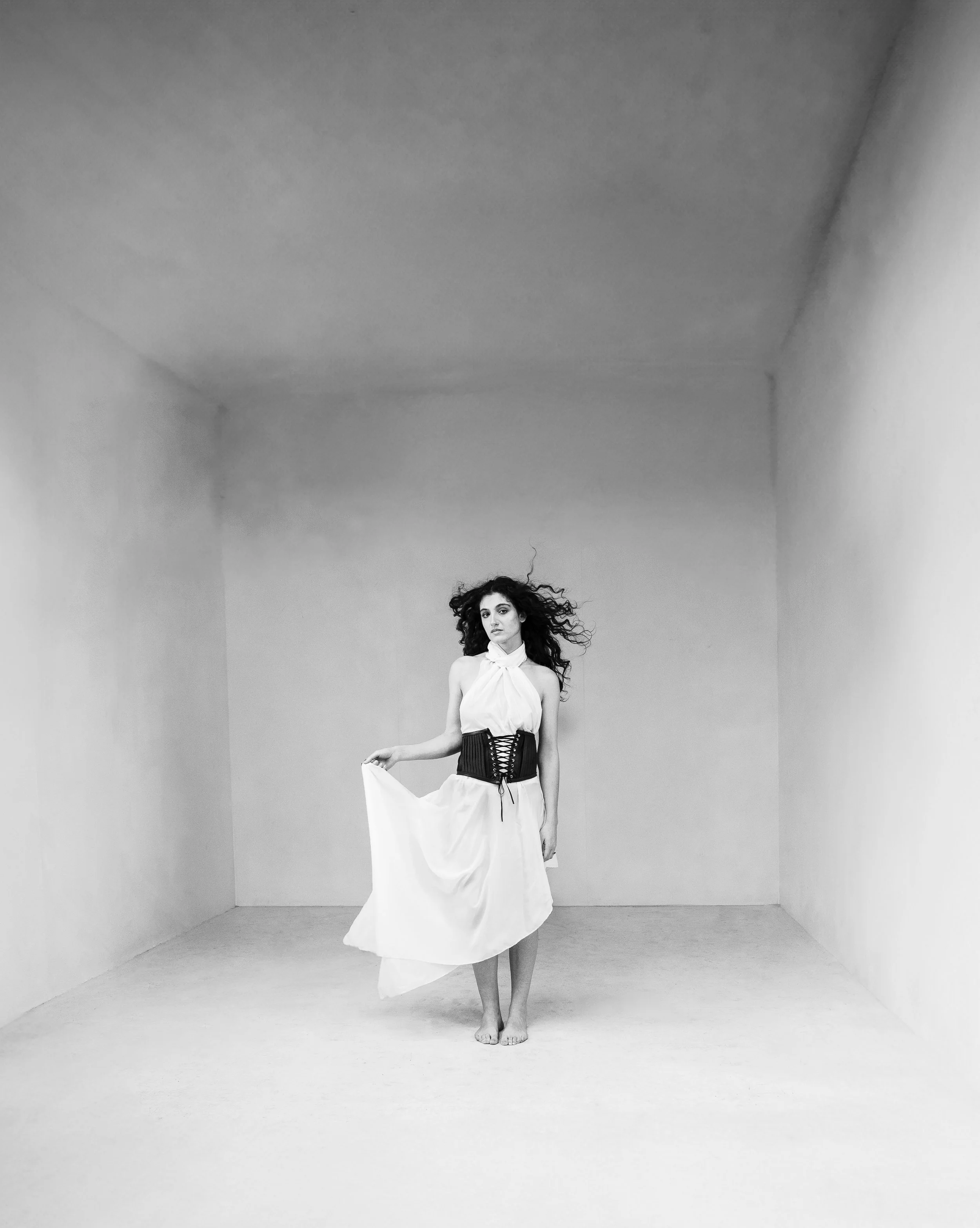 Femme en robe blanche dans une pièce vide avec des murs et un plafond gris clair, cheveux bouclés, portant une ceinture noire corset, tenant un côté de sa robe, regardant vers l'objectif.