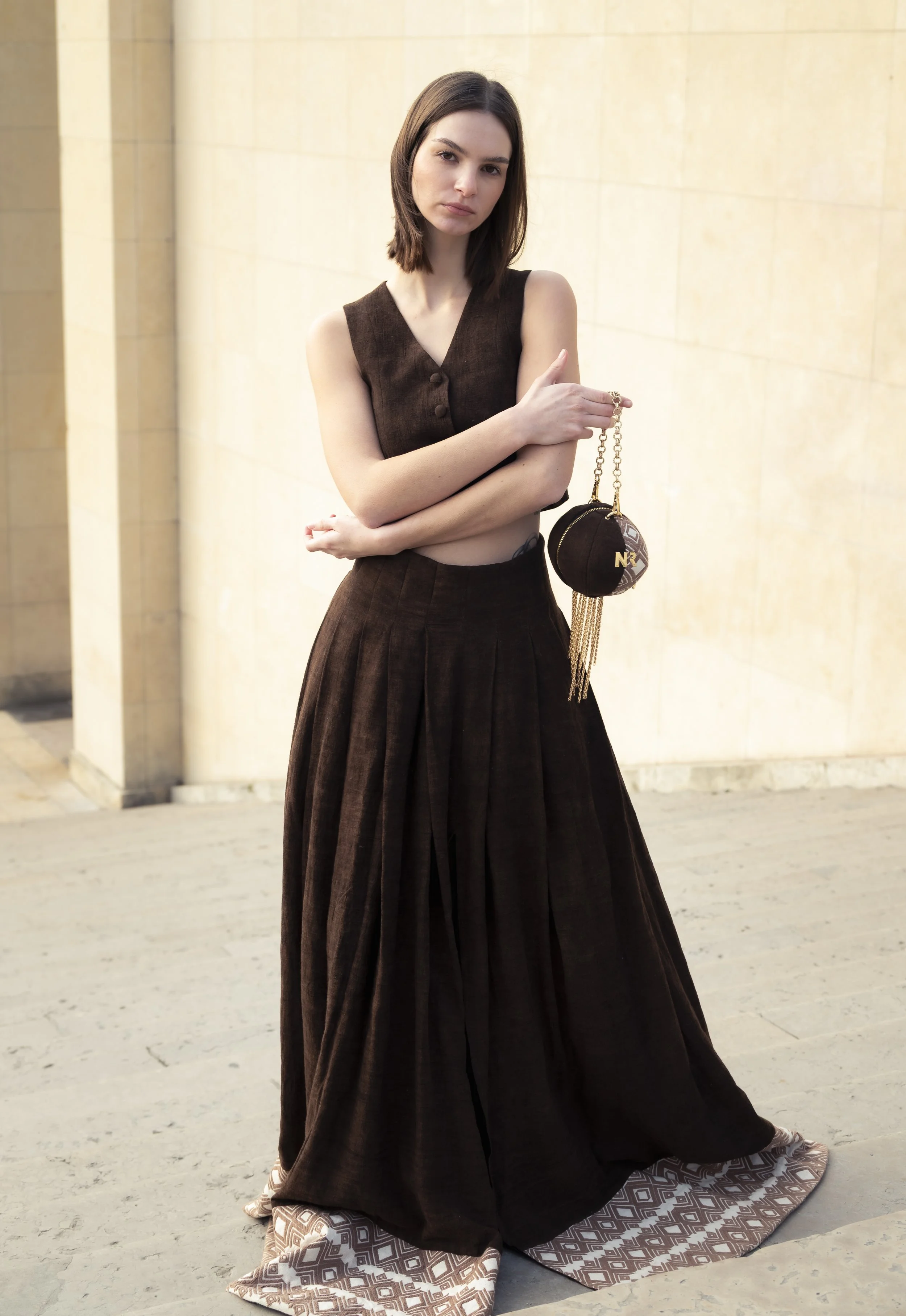 Une femme vêtue d'une jupe longue en tissu marron foncé et d'un haut assorti, tenant un petit sac noir avec une chaîne en or.