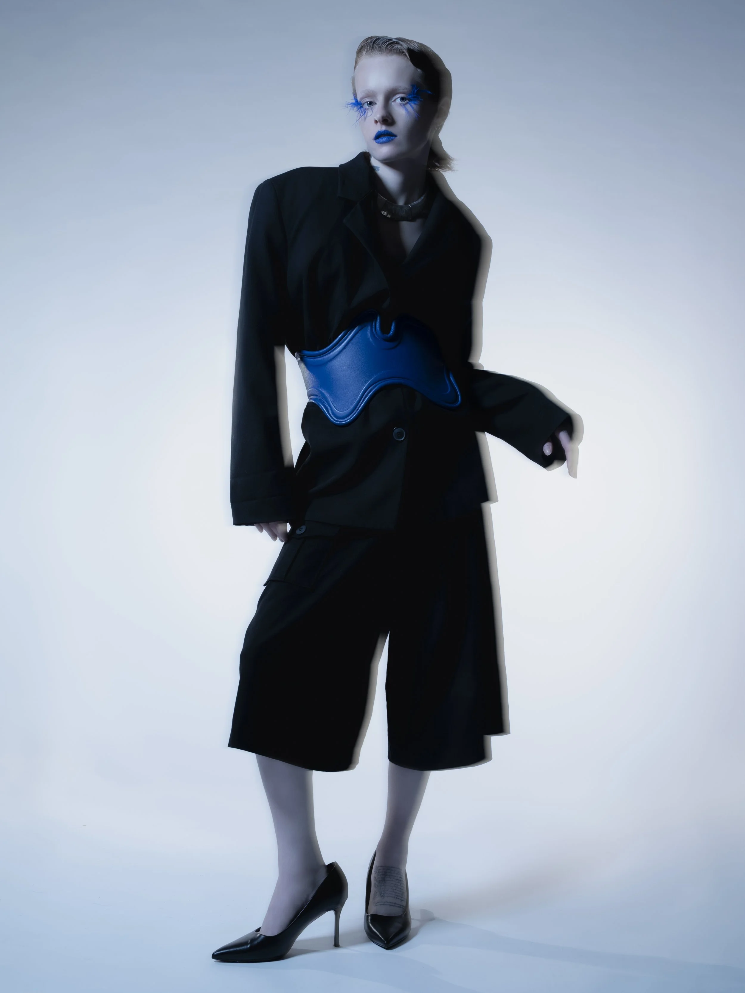 Une femme dans un costume noir avec des chaussures à talons, portant du maquillage bleu vif et des faux cils bleus longs, pose contre un fond clair avec un éclairage dramatique.