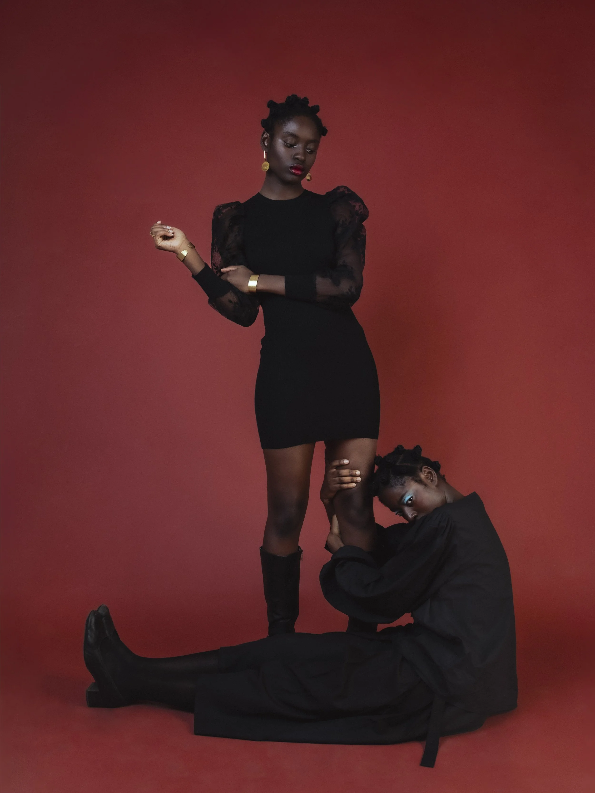 Une femme en robe noire avec des manches en dentelle noire, debout contre un fond rouge, avec une autre femme habillée en noir assise à ses pieds, qui la tient par la jambe. La femme debout a des boucles d'oreilles en or, des bracelets en or, et un r