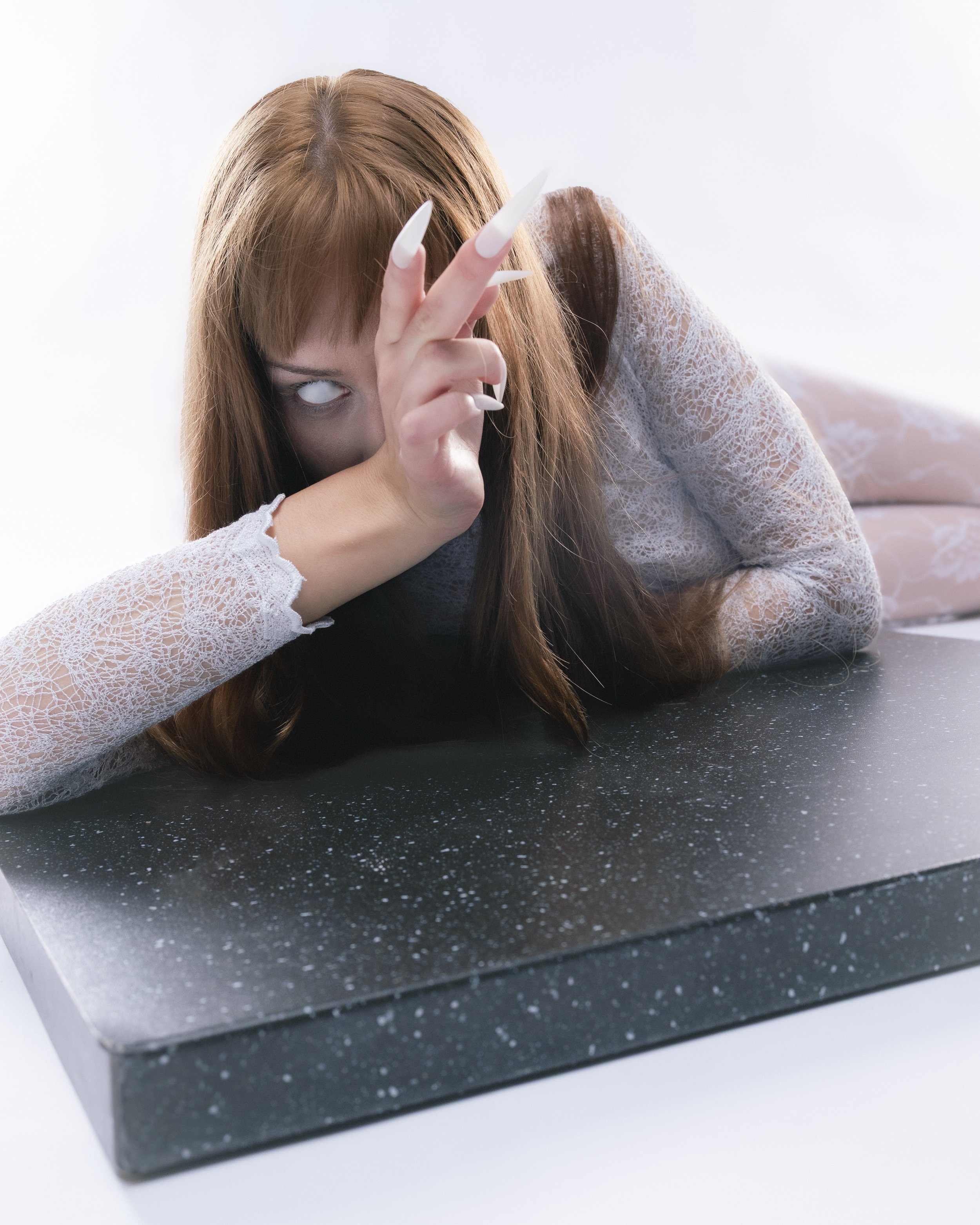 Femme avec longs cheveux roux, maquillage sombre, posant sur une surface noire avec des paillettes, portant un haut en dentelle blanche, faisant un geste mystérieux avec sa main, regardant fixement, dans un contexte sombre et mystérieux.