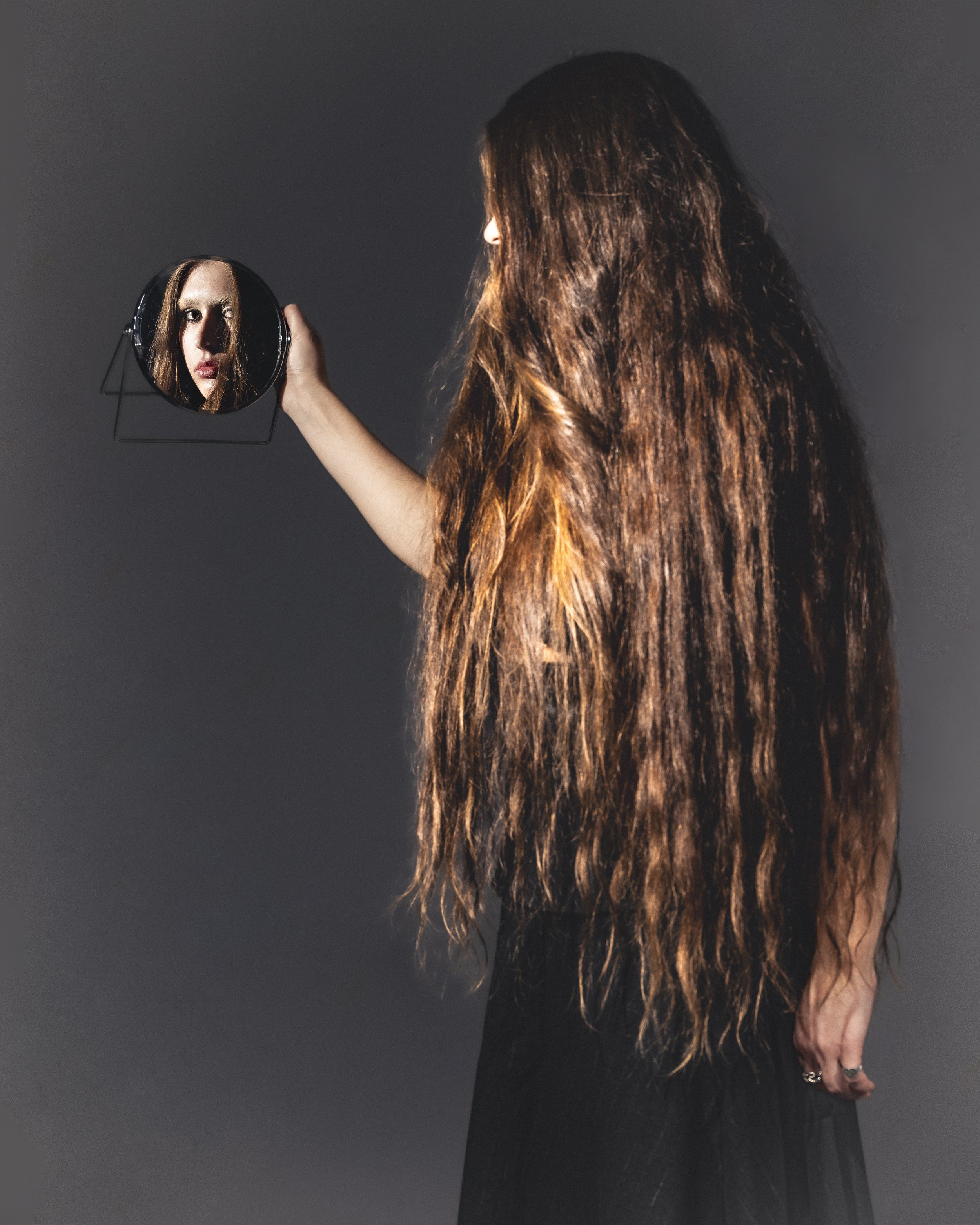 Une femme aux longs cheveux bruns tient un miroir rond, réfléchissant son visage, avec un fond gris