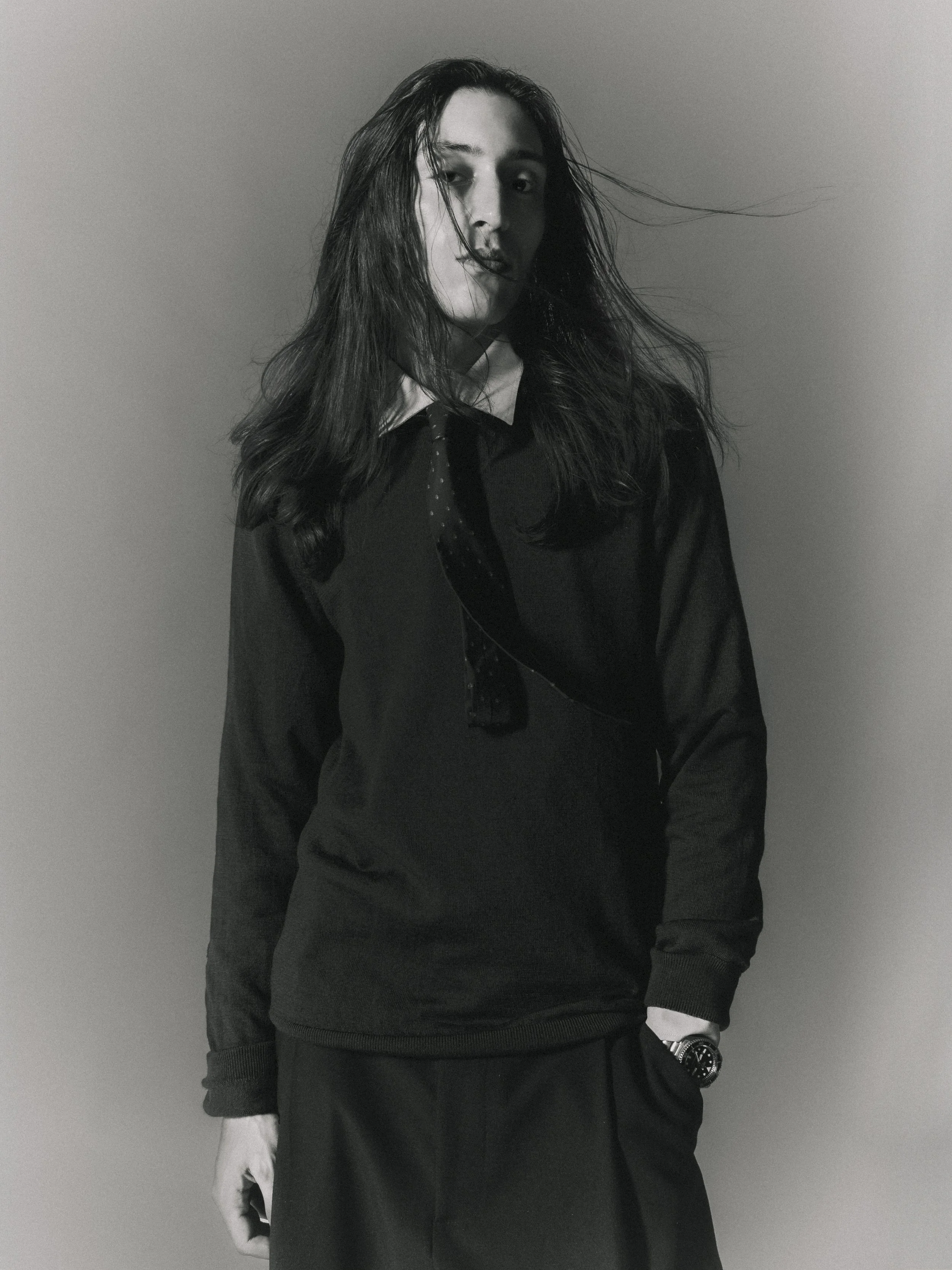 Une femme avec de longs cheveux noirs portant un pull noir, une chemise blanche à col et un nœud papillon noir, posant contre un mur