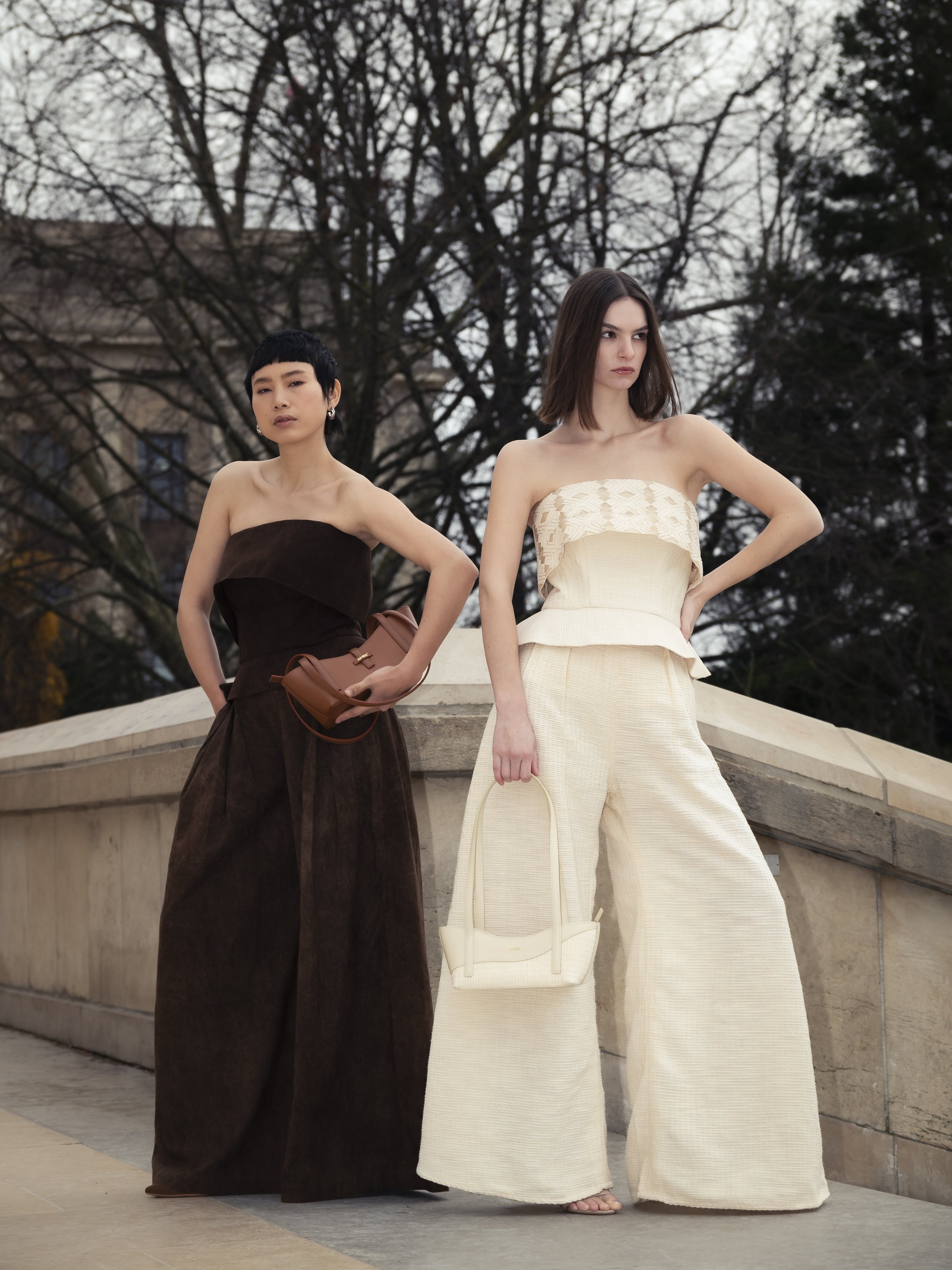 Deux femmes en tenues de mode posent devant une structure urbaine, l'une porte une robe marron et la seconde une tenue beige avec un sac assorti.