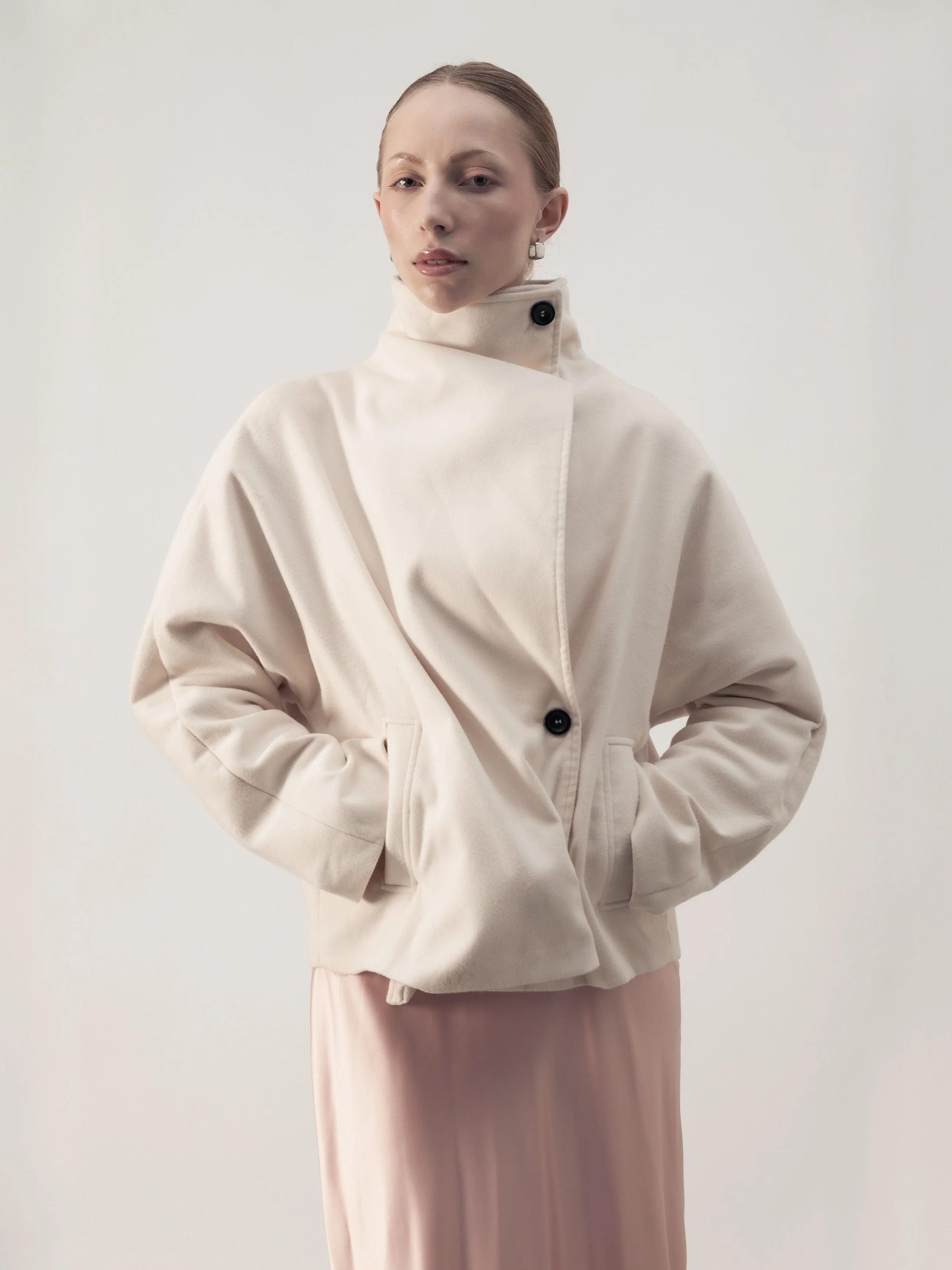 Femme portant un manteau beige avec un col montant et un pantalon rose pâle, posant devant un fond neutre.