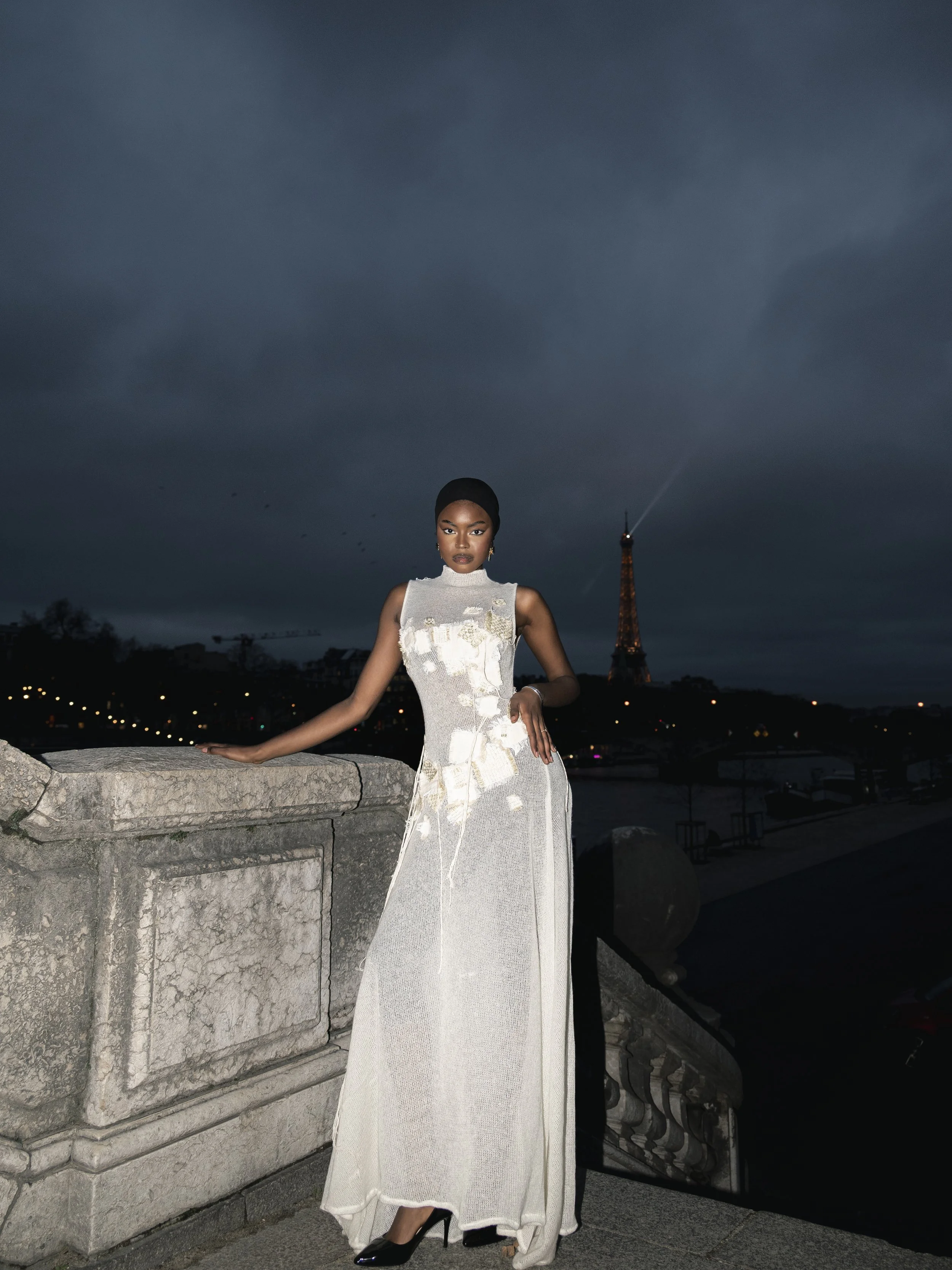 Une femme élégante en robe blanche longue pose devant la Tour Eiffel à Paris, avec un ciel orageux en arrière-plan.