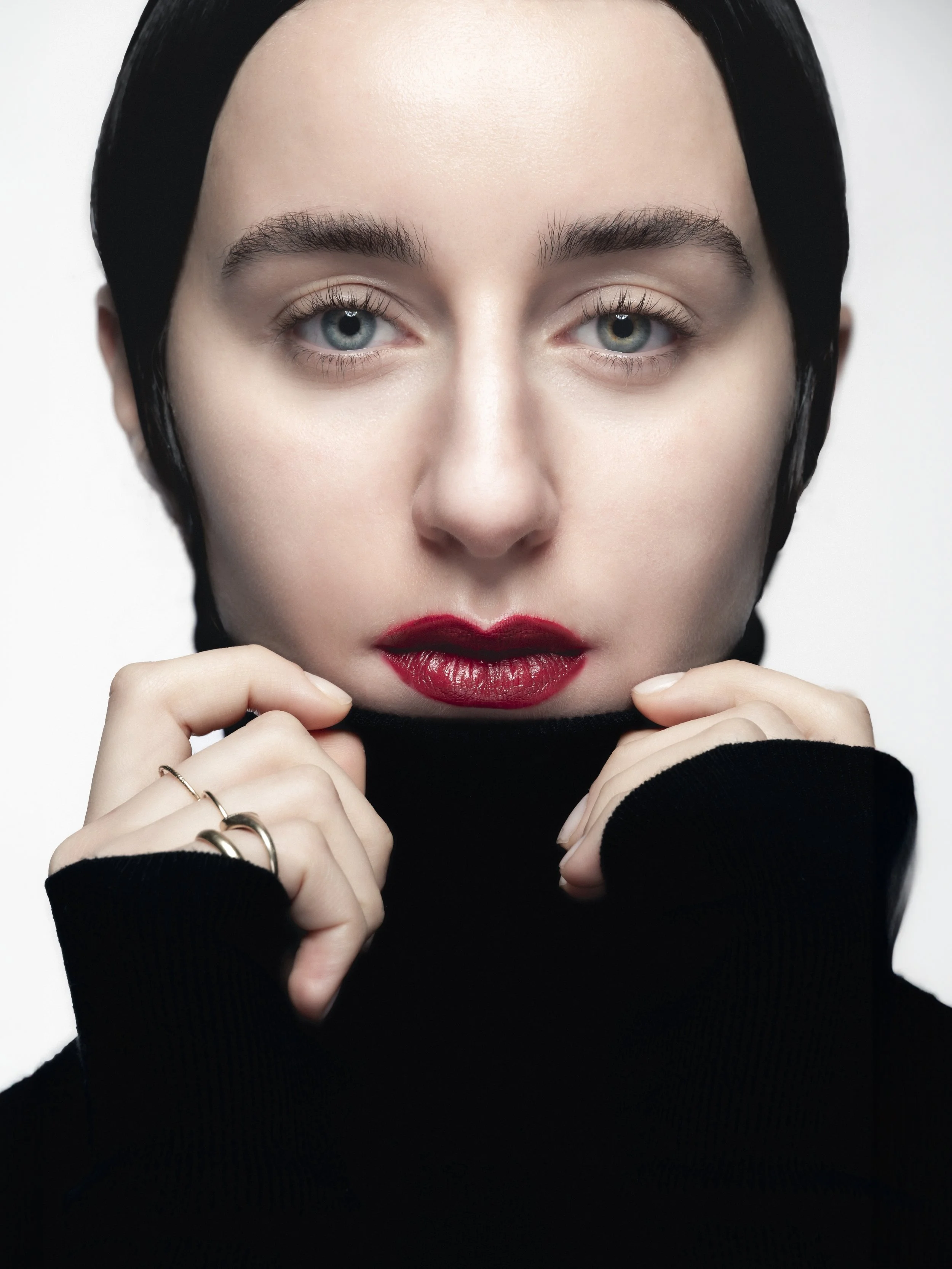 Une femme avec des yeux bleus, des lèvres rouges, portant un pull noir et un chapeau noir, tenant le col de son pull avec ses mains.