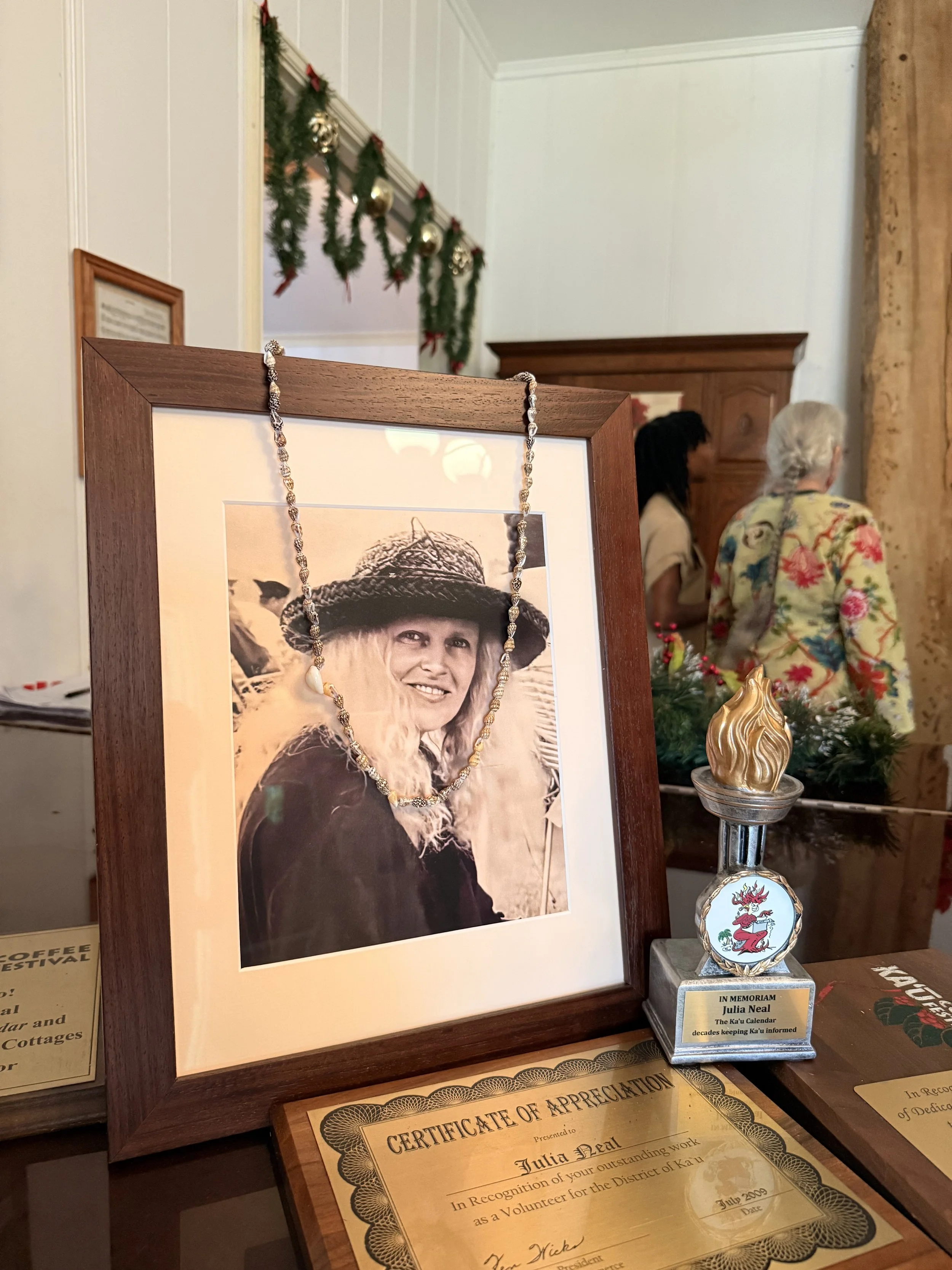 A framed black-and-white photo of a woman wearing a sun hat, with a necklace draped over it, displayed on a table. Behind the table, two women are talking, one wearing a floral sweater and one with dark hair. The setting is decorated for Christmas with garland and ornaments.