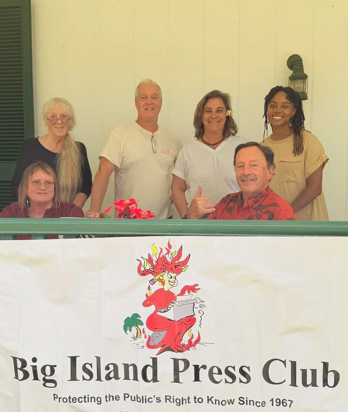 A group of six people, four women and two men, gathered behind a table with a Big Island Press Club banner, with a cartoon goddess illustration, on a porch with cream-colored walls and a lantern on the wall.