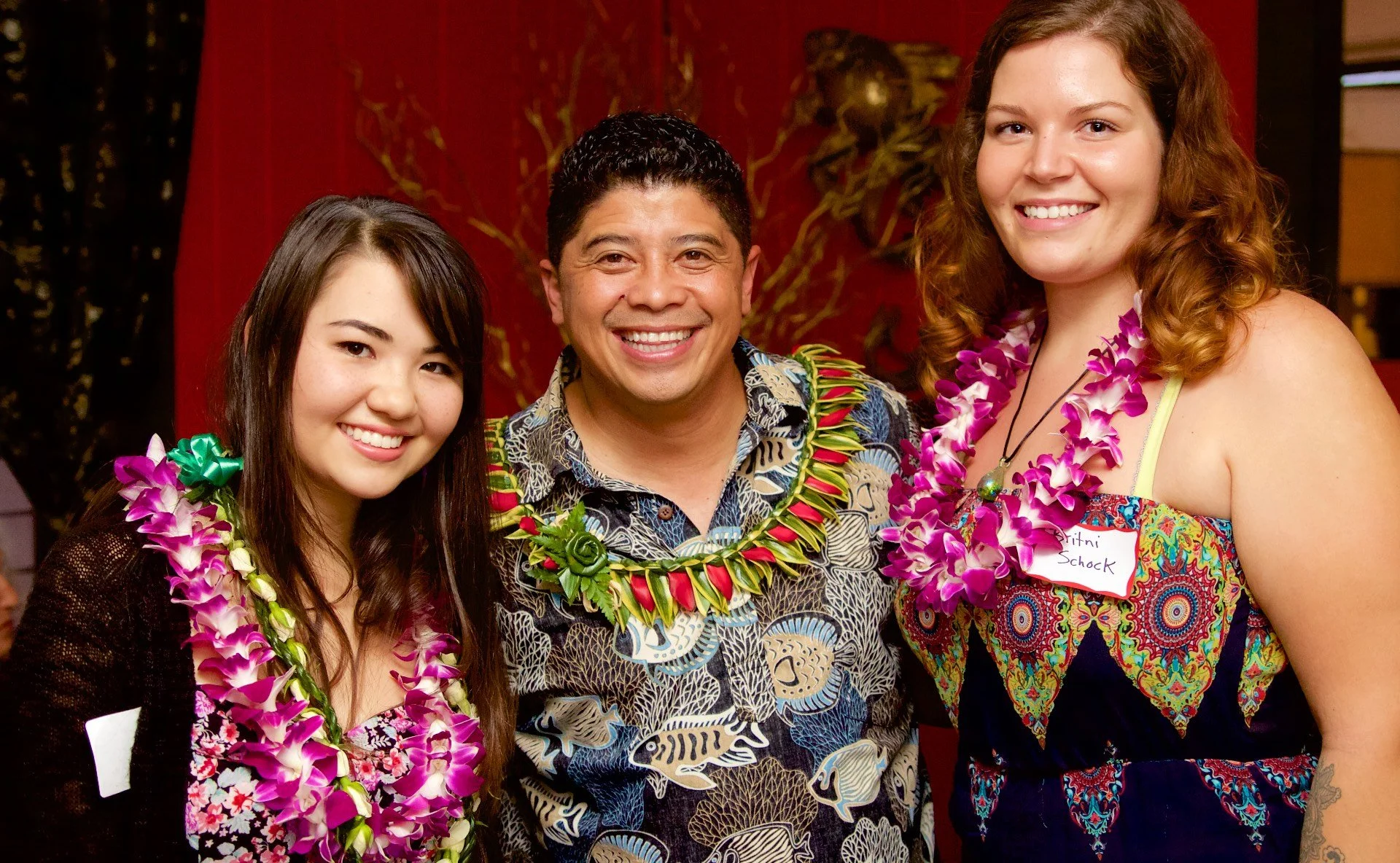 Three people wearing Hawaiian shirts and leis, smiling at a tropical-themed gathering.