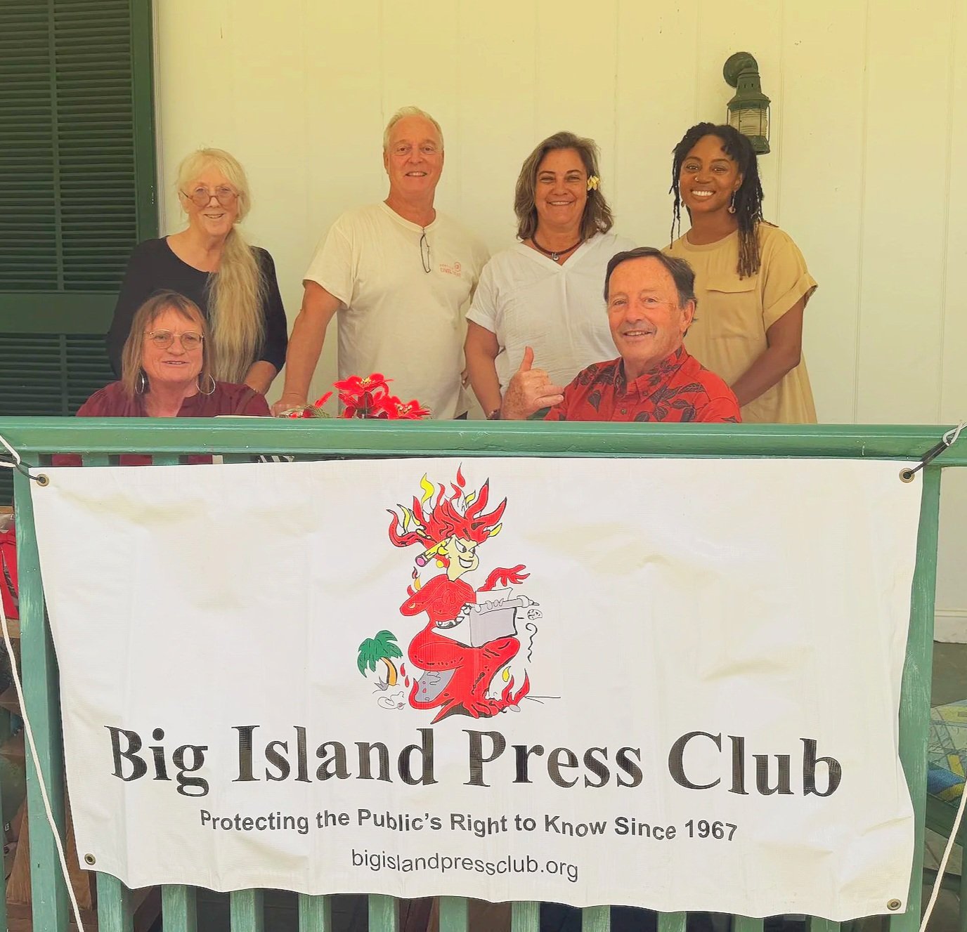 A group of six people, three women and three men, gathered behind a banner that reads 'Big Island Press Club,' in front of a building with a white wall and a lantern. The group is smiling, with one man showing a thumbs-up gesture and a woman with a flower in her hair. The banner features a cartoon fire goddess in red with a typewriter, and text indicating the press club's mission since 1967.