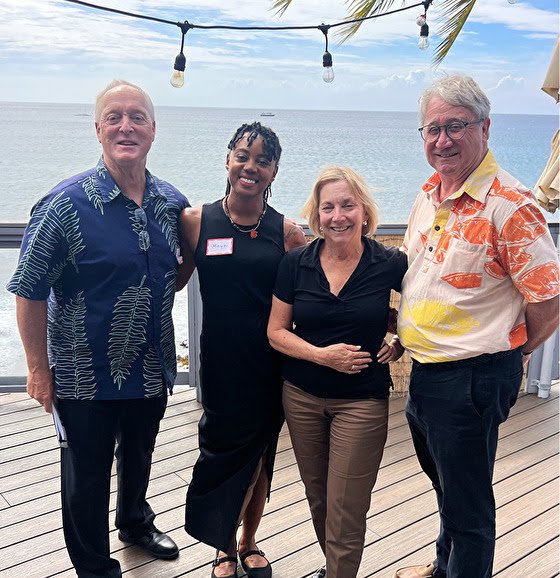 Four people standing on a wooden deck with the ocean and cloudy sky in the background, smiling for the photo.
