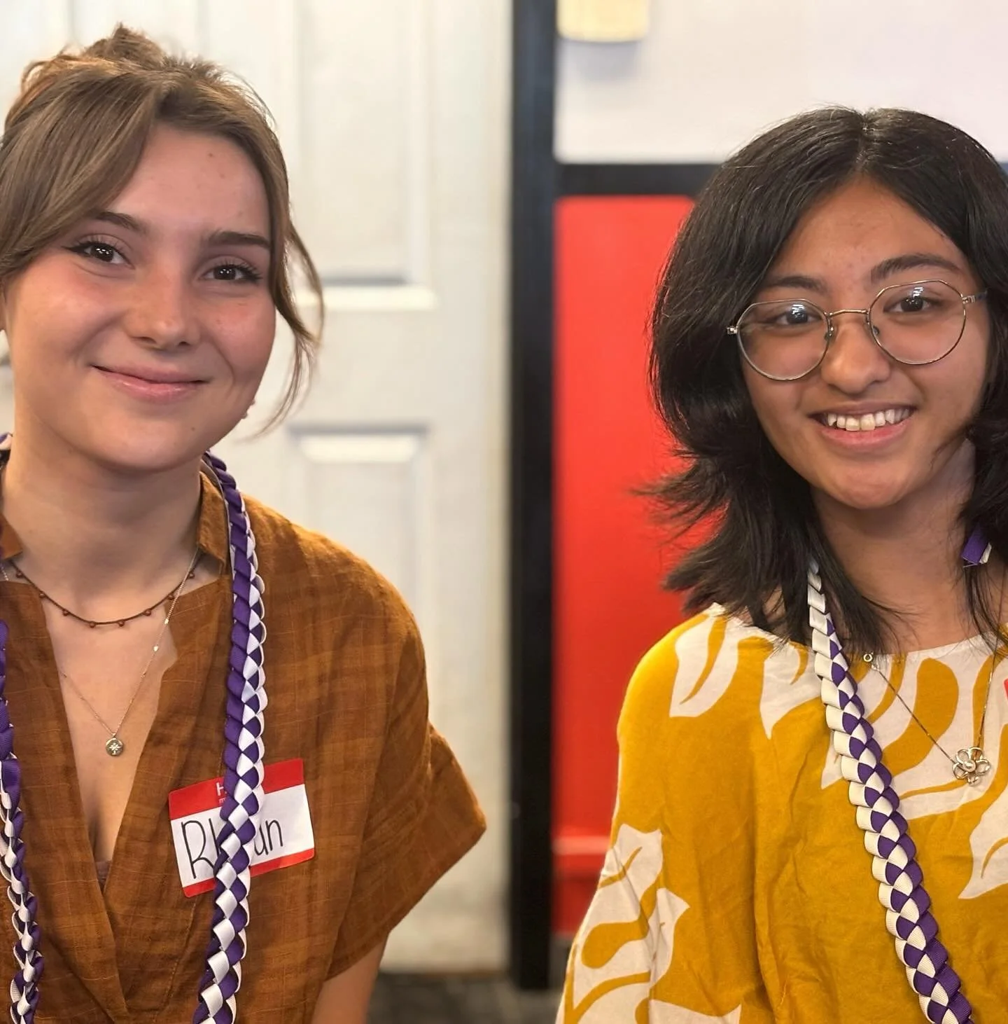 Two women smiling at an event, wearing purple and white leis around their necks. The woman on the left has short, light brown hair, wearing an orange shirt with a name tag, and is wearing two layered necklaces. The woman on the right has medium-length dark hair, wearing glasses, and a yellow and white patterned top.