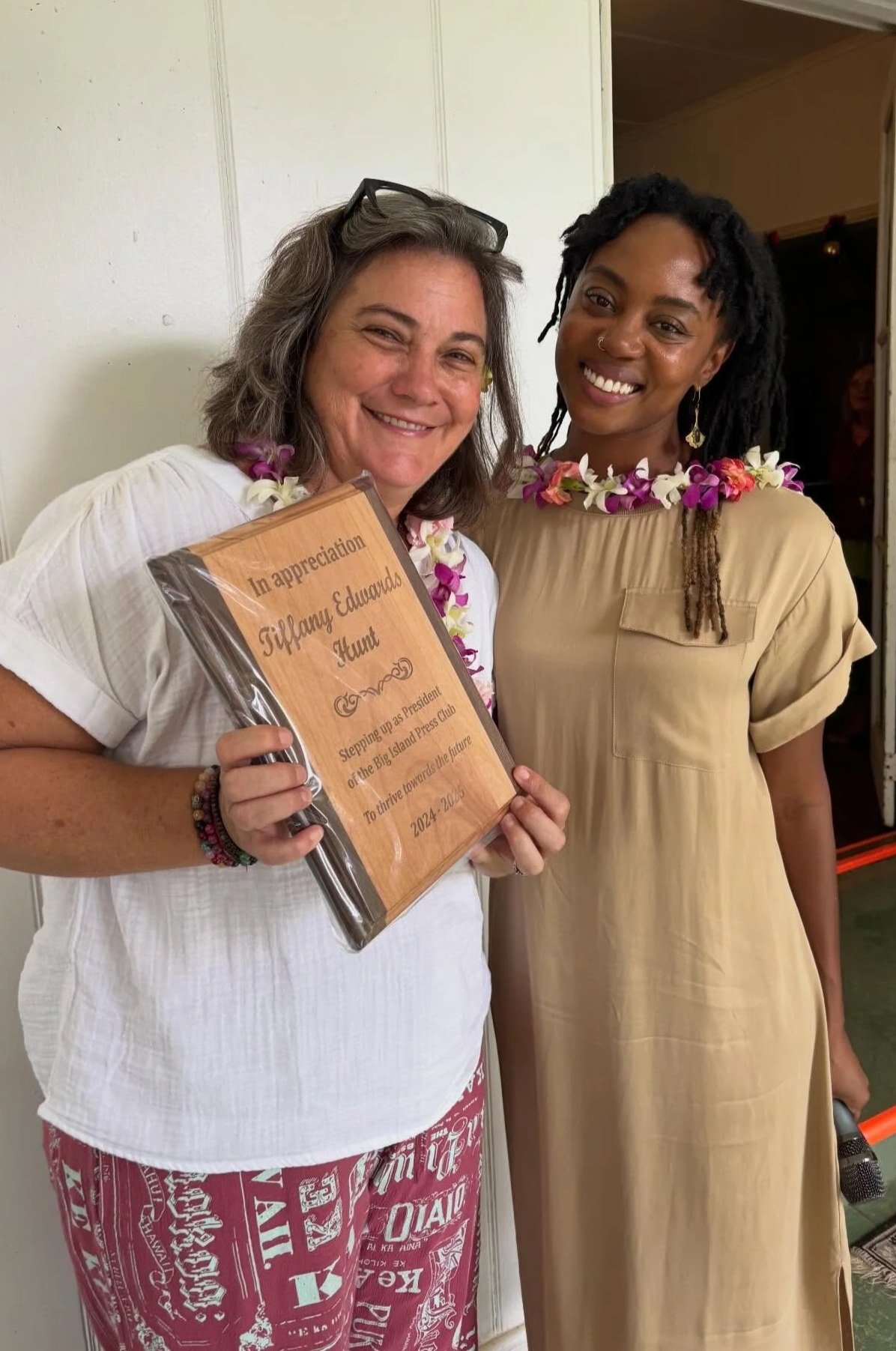 Two women wearing leis pose together indoors. One woman holds a wooden plaque with an appreciation message. Both are smiling.