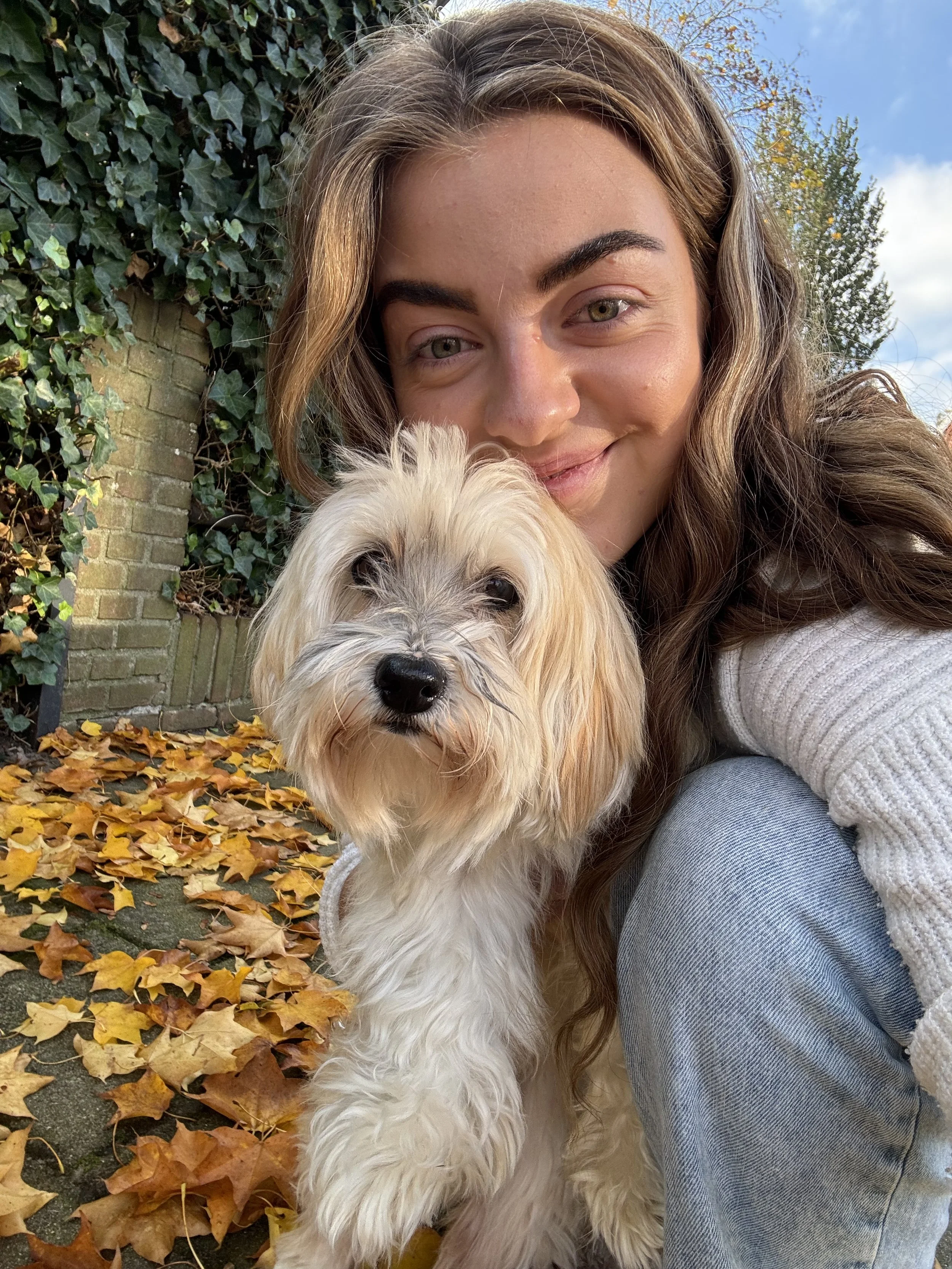Een jonge vrouw met lang, bruin haar neemt een selfie met haar lichtbruine en witteukelige hond op een herfstige dag, omgeven door gele en oranje bladeren onder een blauwe lucht.