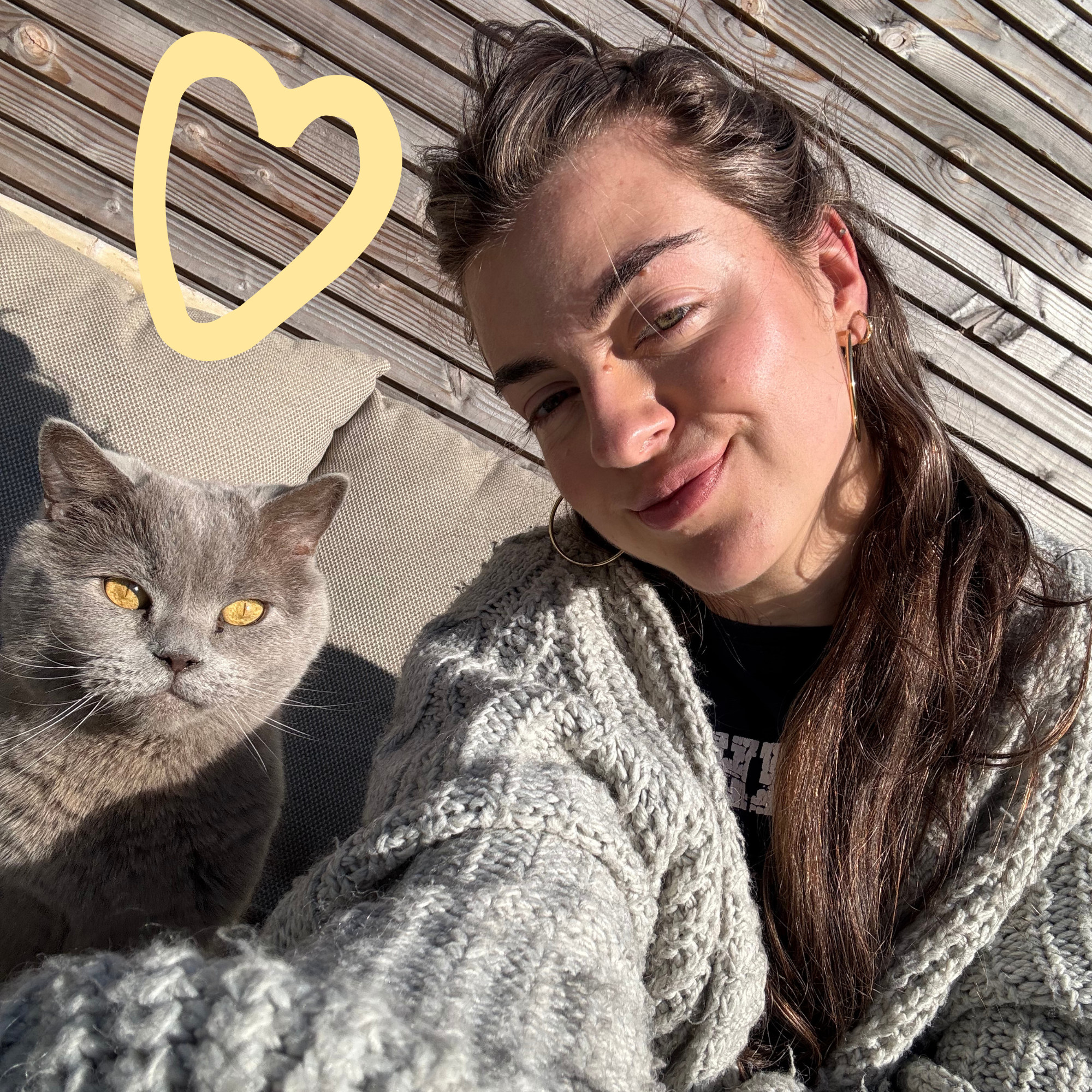 Een vrouw die een selfie neemt met haar kat op een buitenbank onder de zon, met een houten achtergrond en een geel hart op de foto.