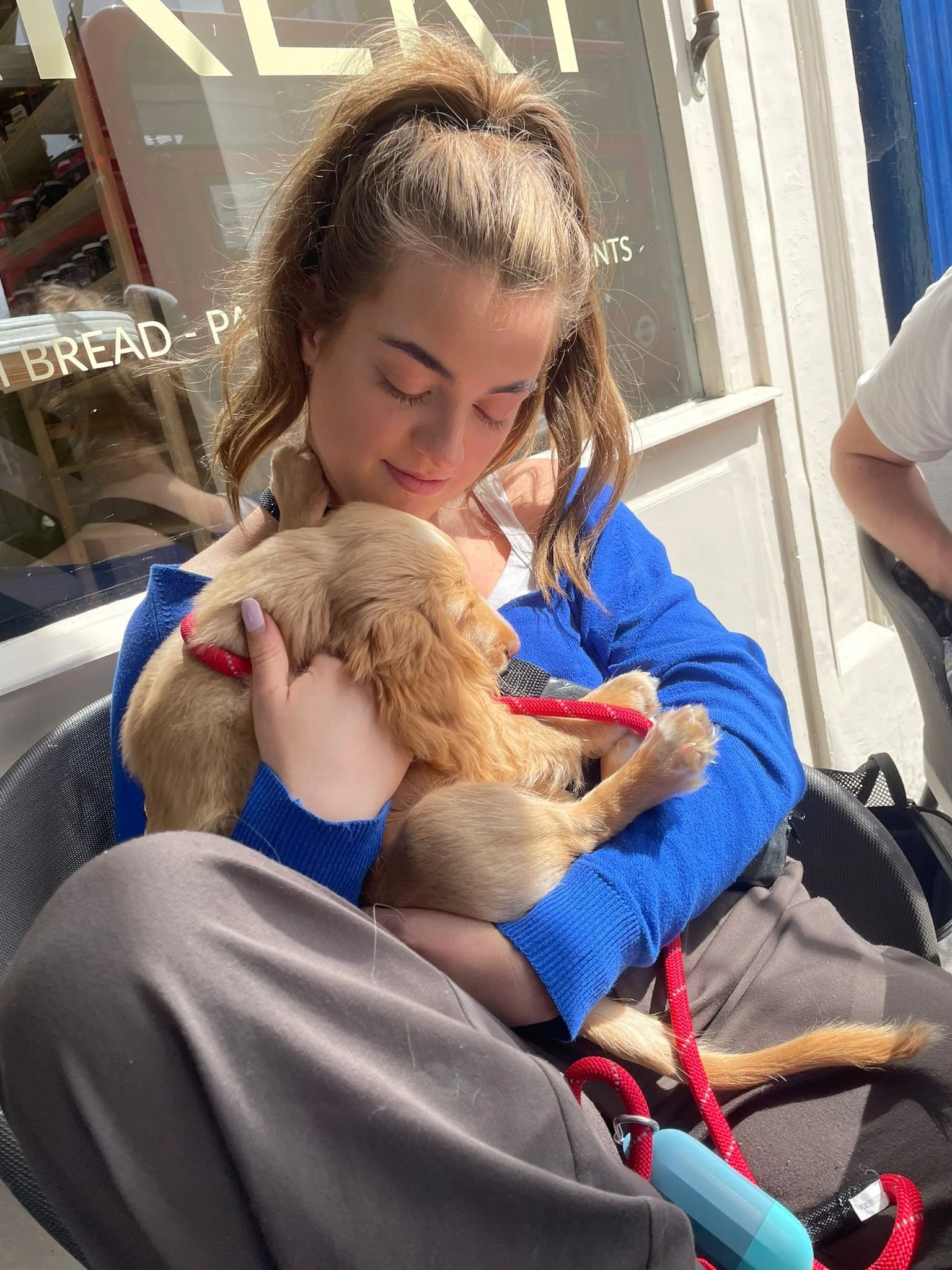 Een jonge vrouw met bruin haar in een hoge paardenstaart die een klein golden retriever puppy vasthoudt en likt. Ze zitten buiten bij een café of bakkerij met een raam en tekst op de achtergrond.