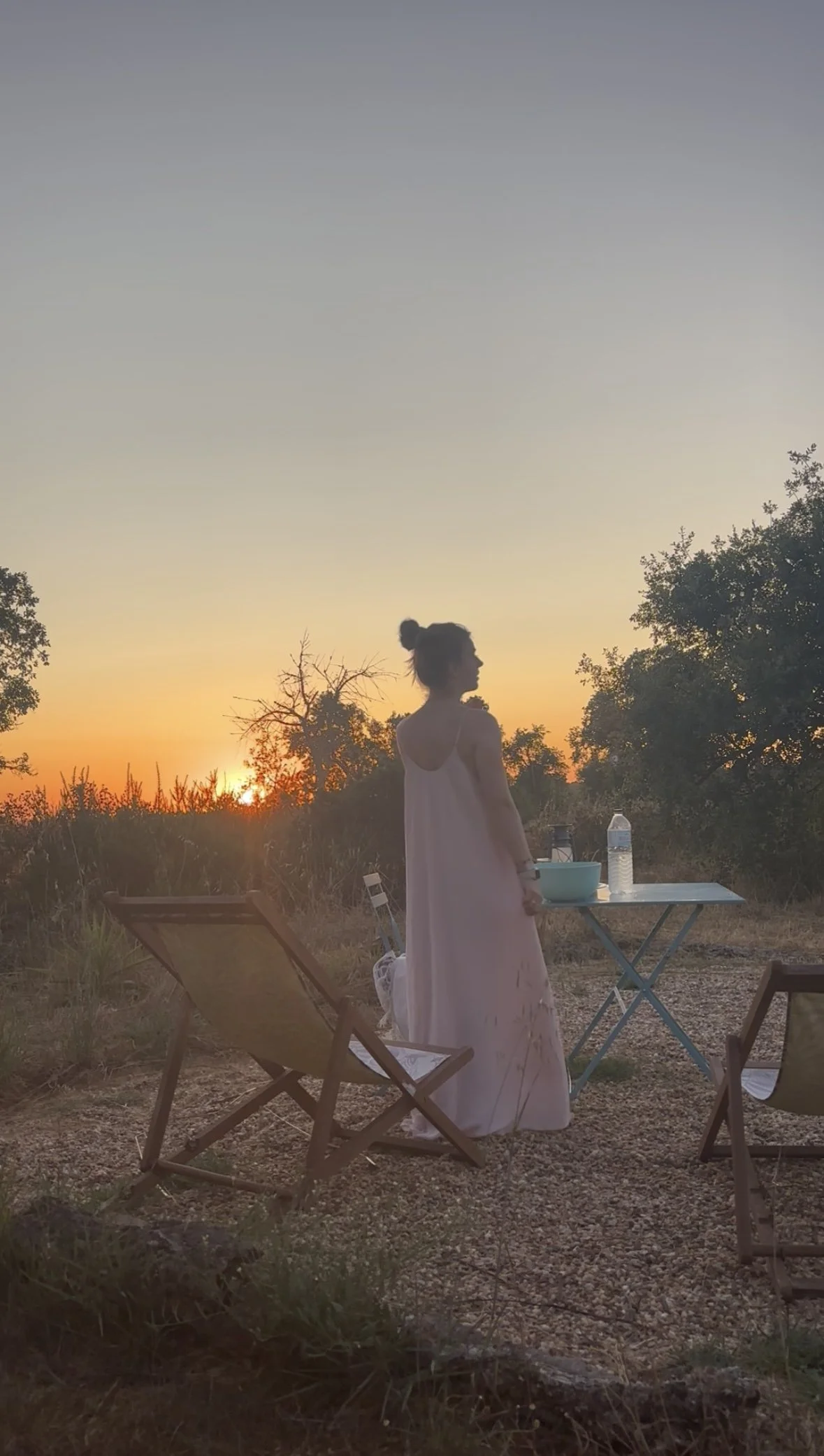 Een vrouw in een lange witte jurk staat in een natuurlijk landschap tijdens zonsondergang, met twee klapstoelen en een tafel met water en een kom.