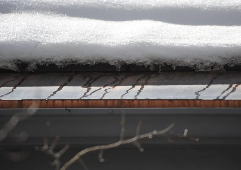 Close-up of a snow-covered window ledge with snow on top and ice forming on the edge, below a wooden window frame.