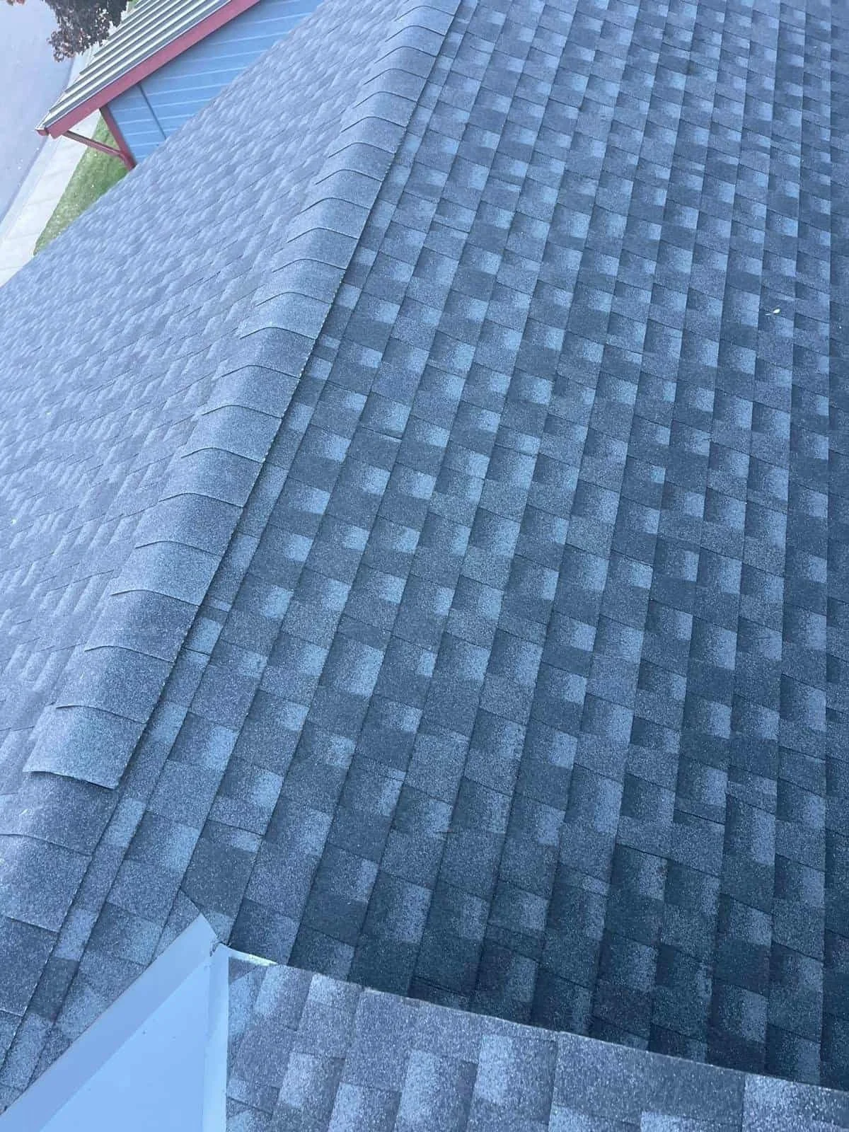A close-up view of a dark gray asphalt shingle roof on a residential building, with a part of the white wall visible in the foreground.