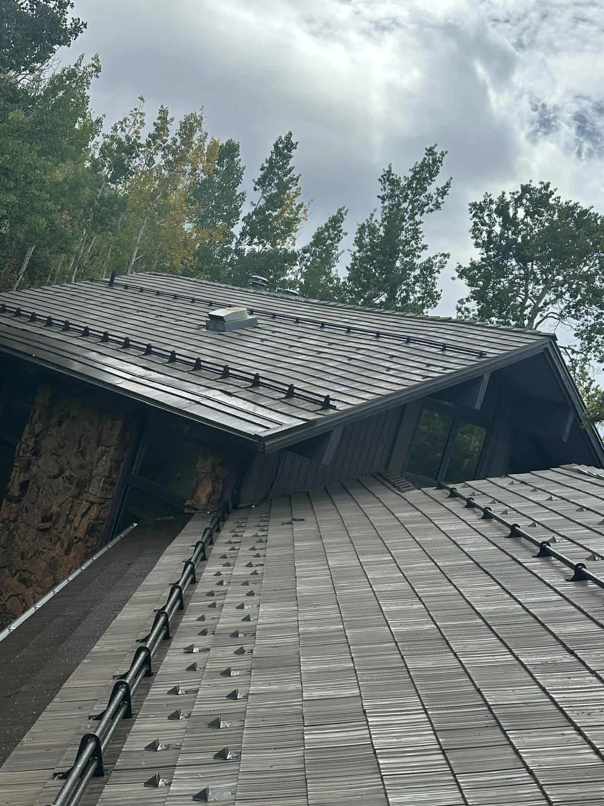 A roof with metal panels under construction, with visible fasteners and black roof snow guards, surrounded by tall trees and a cloudy sky.