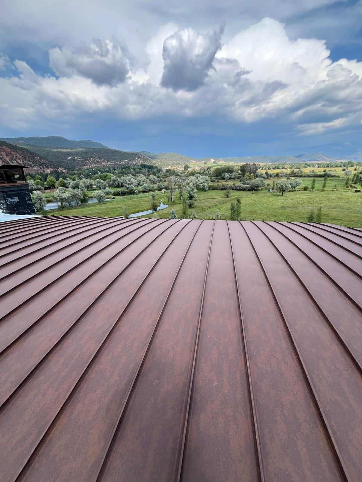 Green fields with trees, a small river, and distant mountains under a blue sky with large clouds, viewed from a rooftop with brown metal panels.